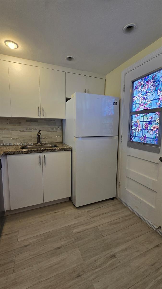 Kitchenette featuring white shaker-style cabinetry, stone-surface countertops, textured tile backsplash, wood-finish flooring, and a decorative stained-glass panel door