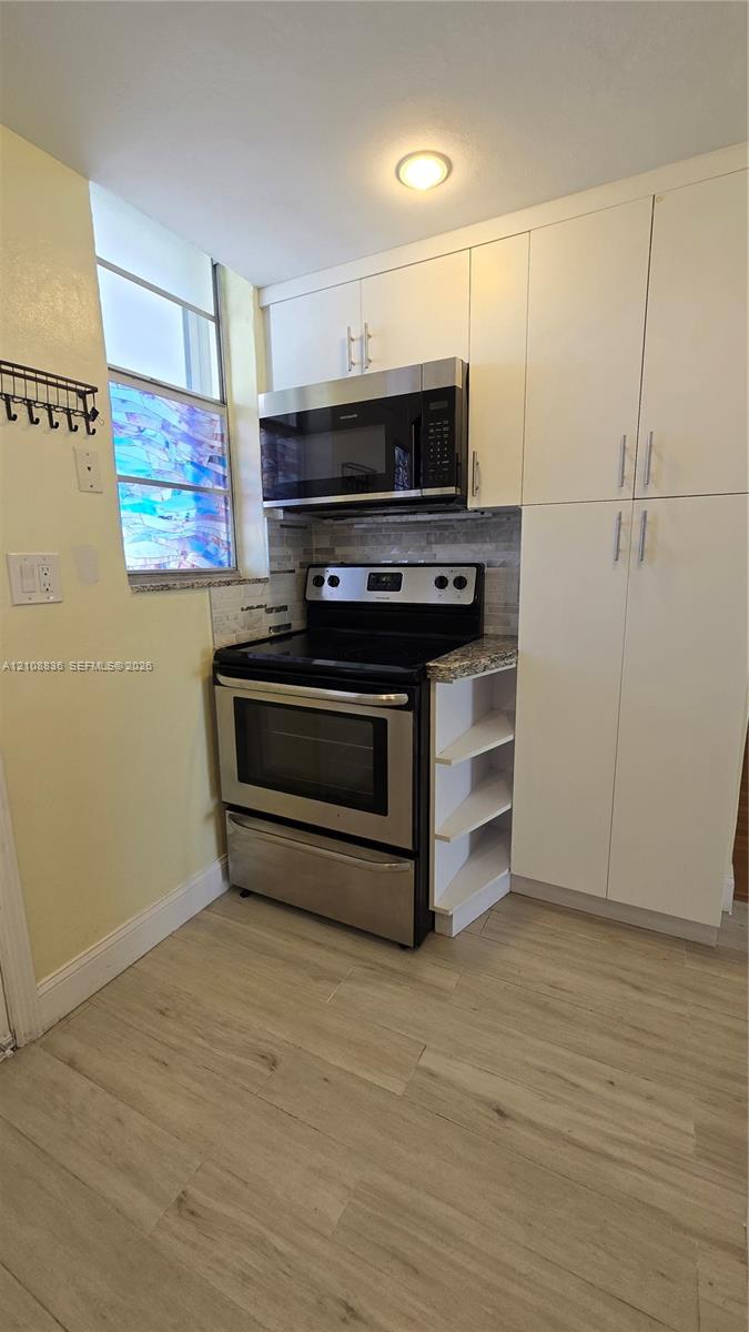 Kitchen featuring white cabinetry with brushed-nickel hardware, stainless steel appliances, stone-look countertops, tile backsplash, and light wood-finish flooring