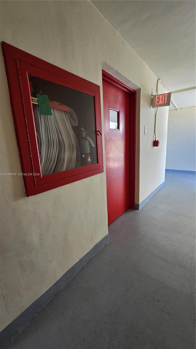 Hallway featuring a painted metal door, wall-mounted fire safety cabinet with glass panel, and illuminated exit signage