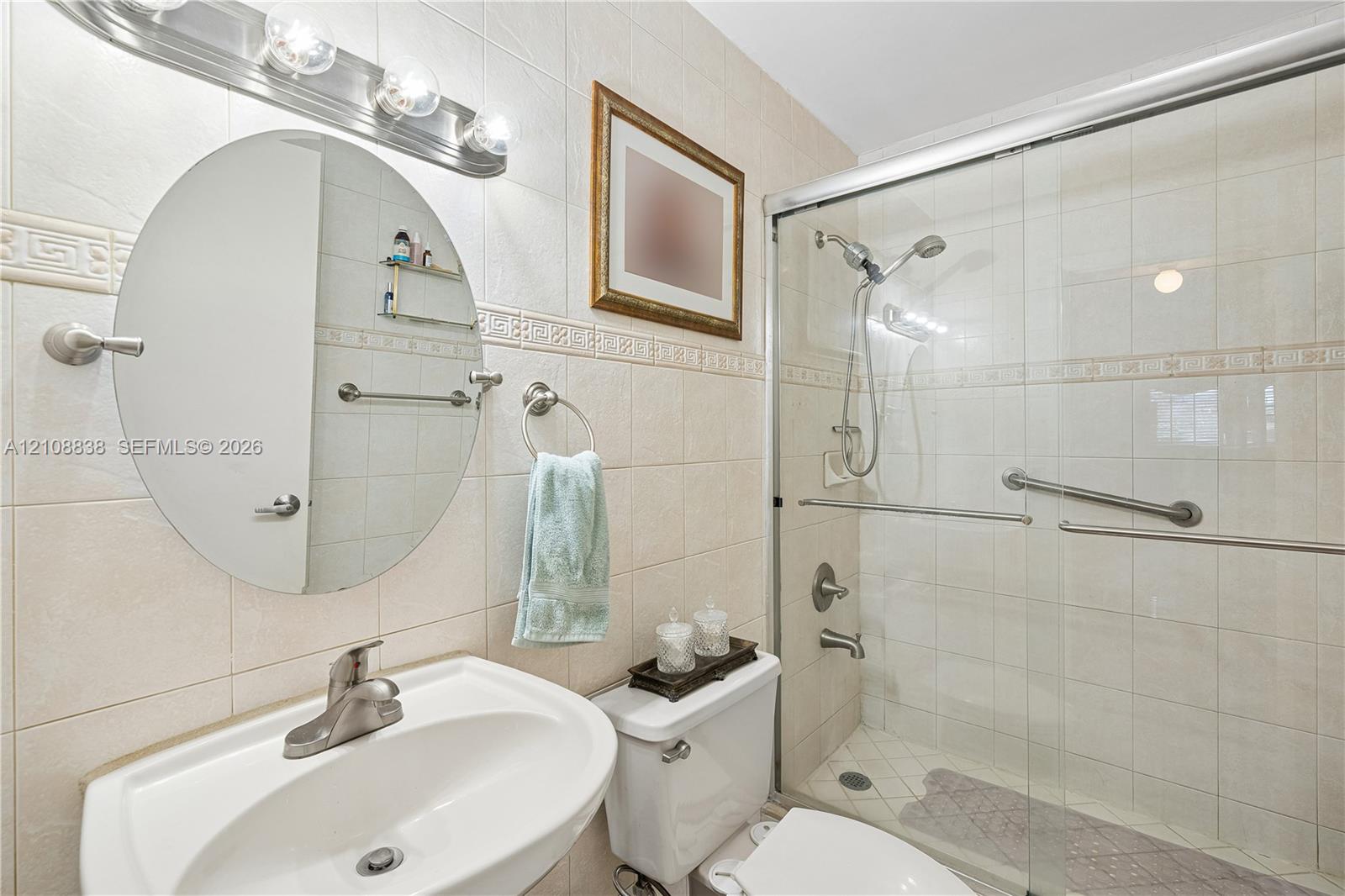 Bathroom featuring ceramic wall tiling with decorative trim, oval vanity mirror, brushed-nickel hardware, and a walk-in shower enclosure with a sliding glass door
