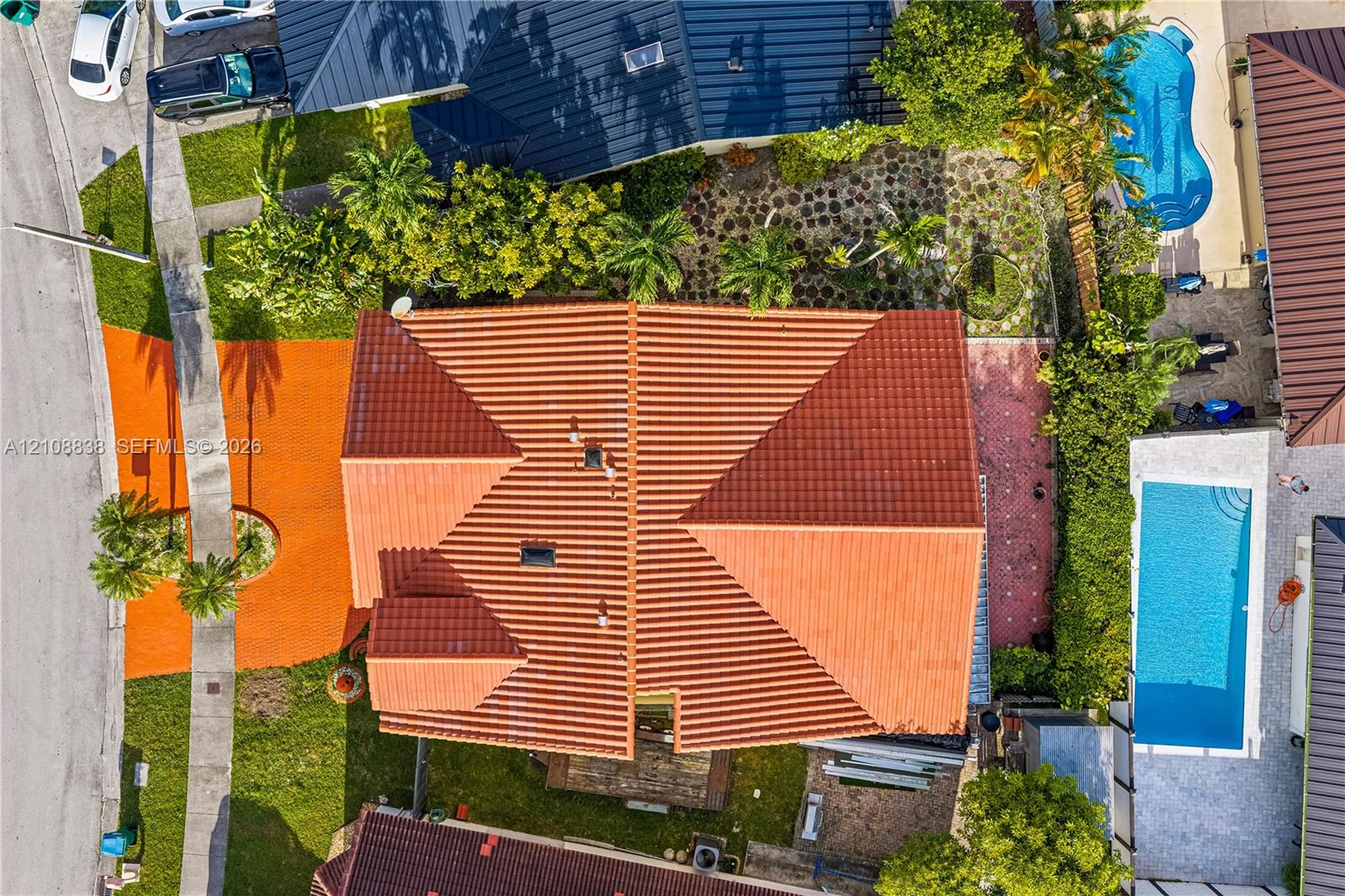 Aerial view of a property featuring a multi-pitched clay tile roof, a private swimming pool, landscaped lawn, and decorative stone paving