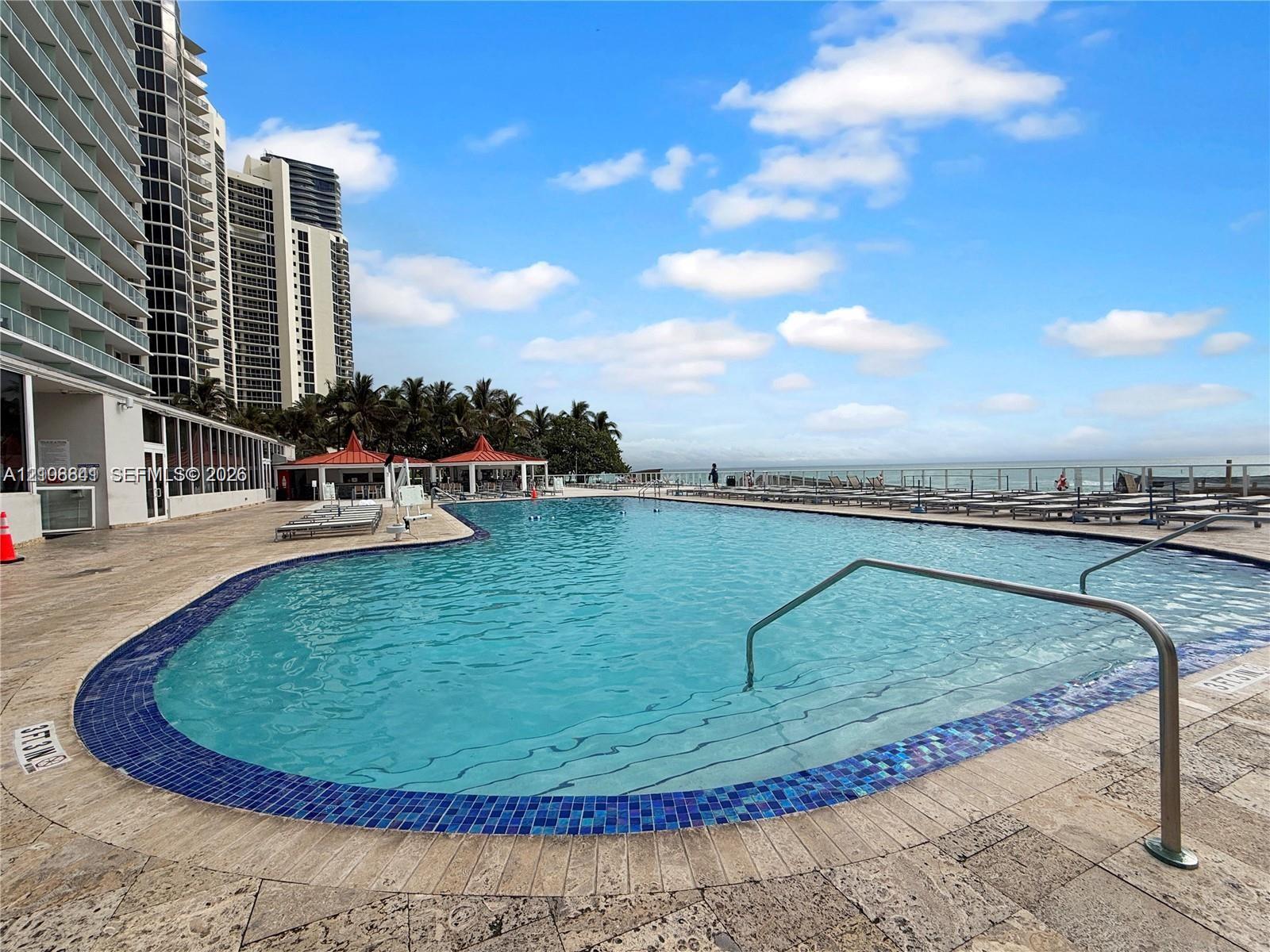 Curved swimming pool featuring blue tile coping, stainless steel handrails, and a natural stone deck