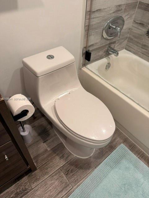 One-piece elongated toilet featuring a dual-flush button, wood-finish tile flooring, and a bathtub surround with grey-toned textured tile and chrome fixtures