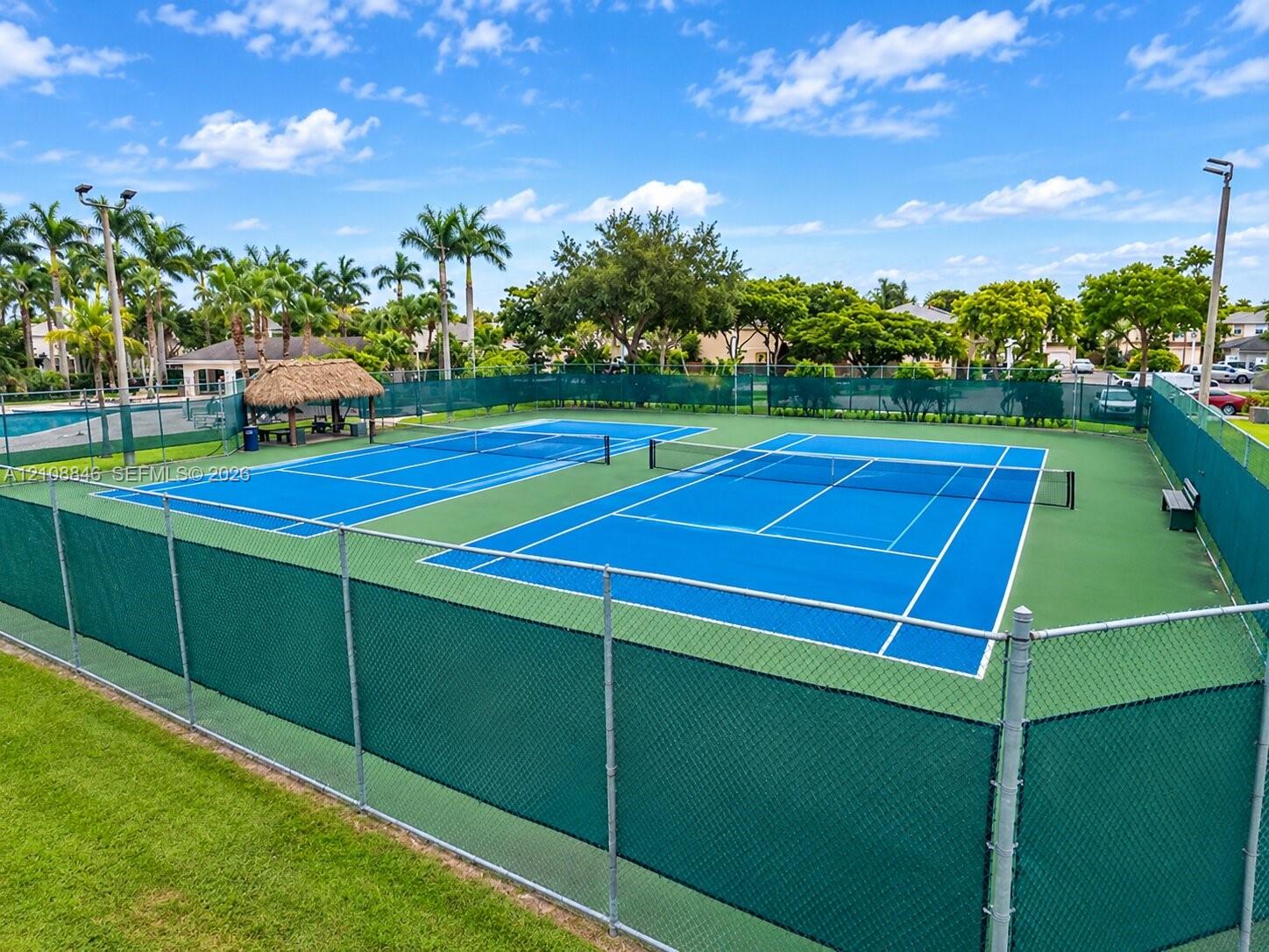 Dual tennis courts featuring high-tensile chain-link fencing, a thatched-roof pavilion, and integrated court lighting