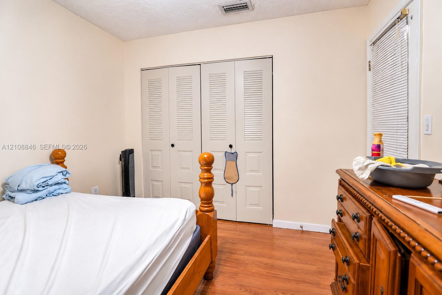 Wood-finish flooring, dual-panel closet doors with louvered upper sections, and a window fitted with horizontal blinds