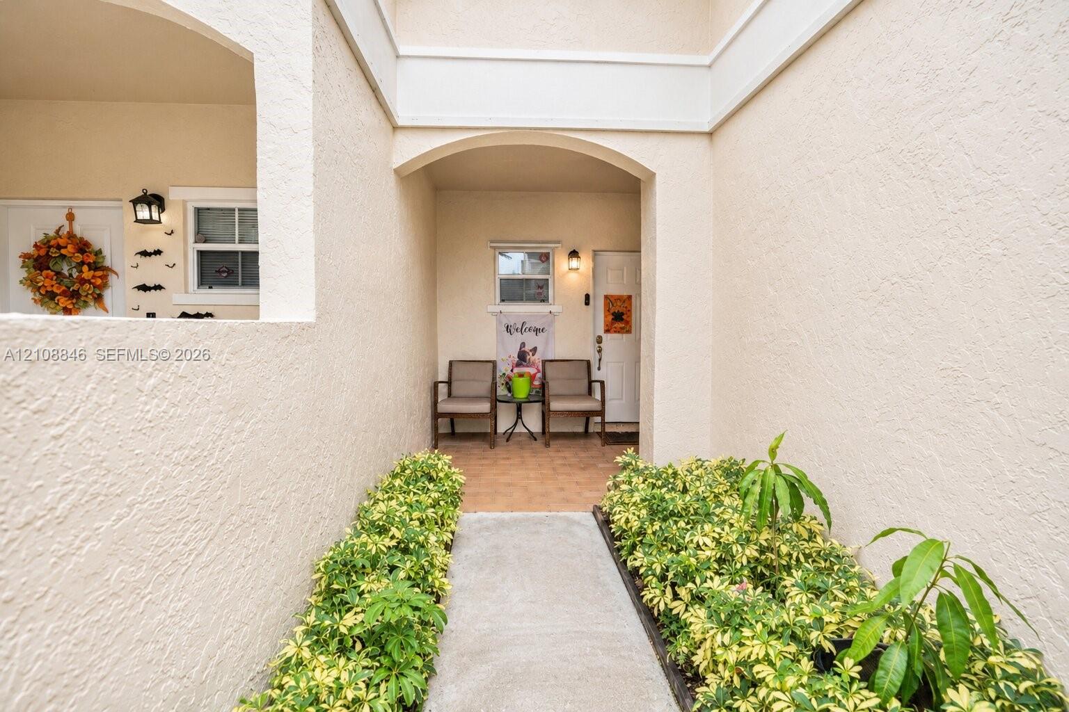 Stucco exterior entryway featuring a concrete walkway, arched architectural detailing, wall-mounted light fixtures, and manicured perimeter landscaping