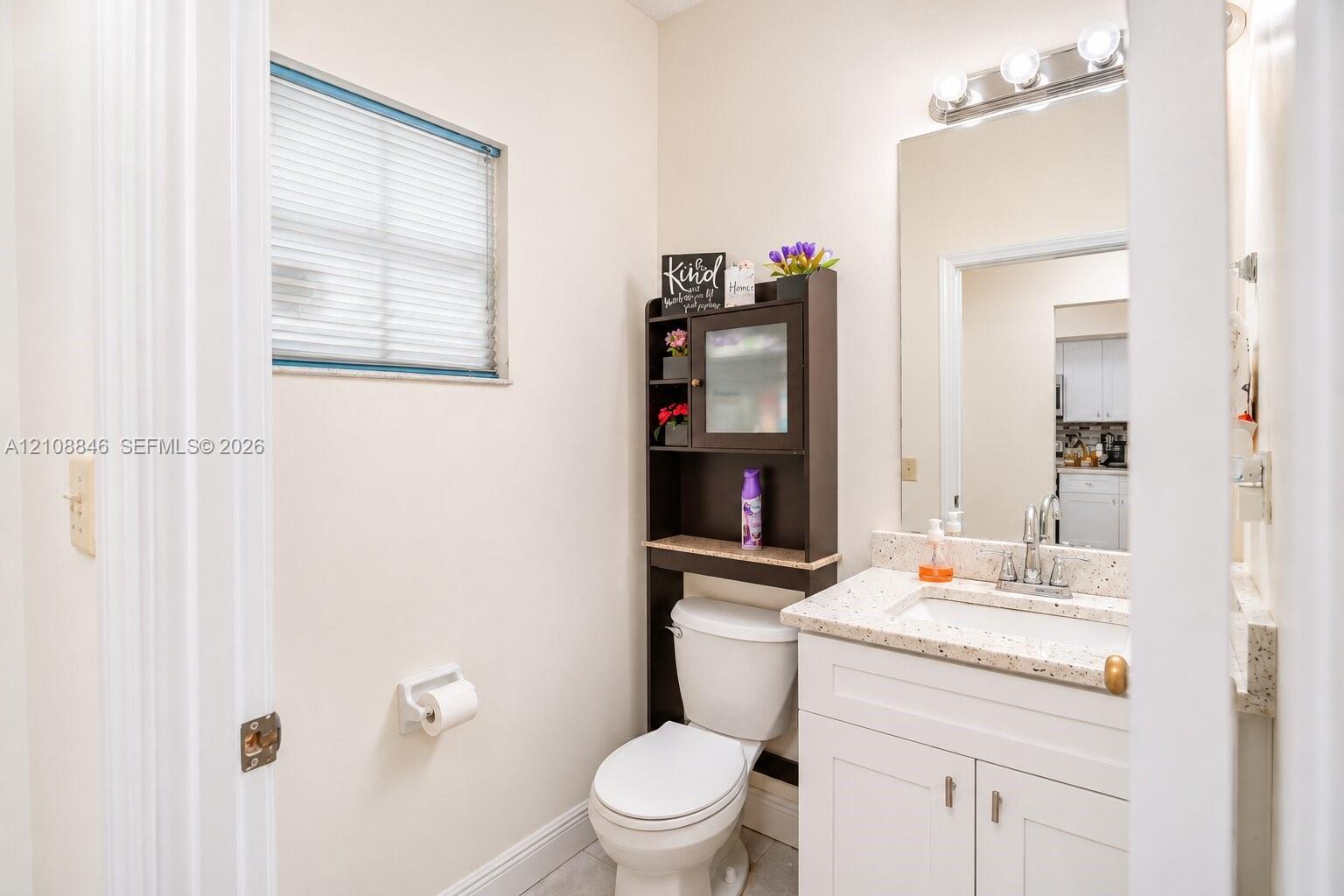 Bathroom featuring a vanity with a white cabinet and stone-composite countertop, integrated sink, wall-mounted mirror, four-bulb light fixture, and horizontal blind window