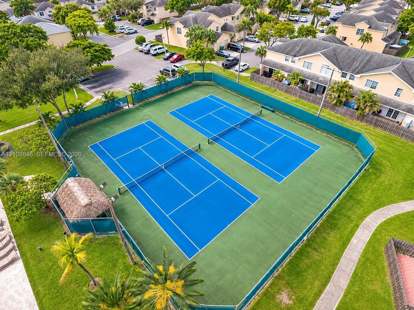 Dual tennis courts featuring hard-surface playing areas, perimeter fencing, and an integrated thatched-roof structure