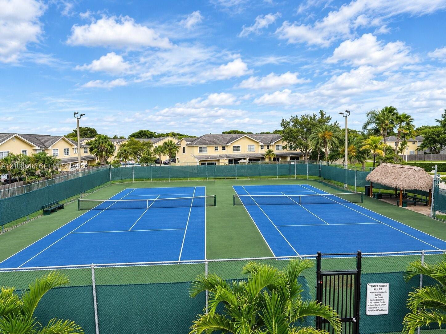 Dual tennis courts featuring specialized surface markings, perimeter chain-link fencing, stadium-style lighting, and a thatched-roof pavilion