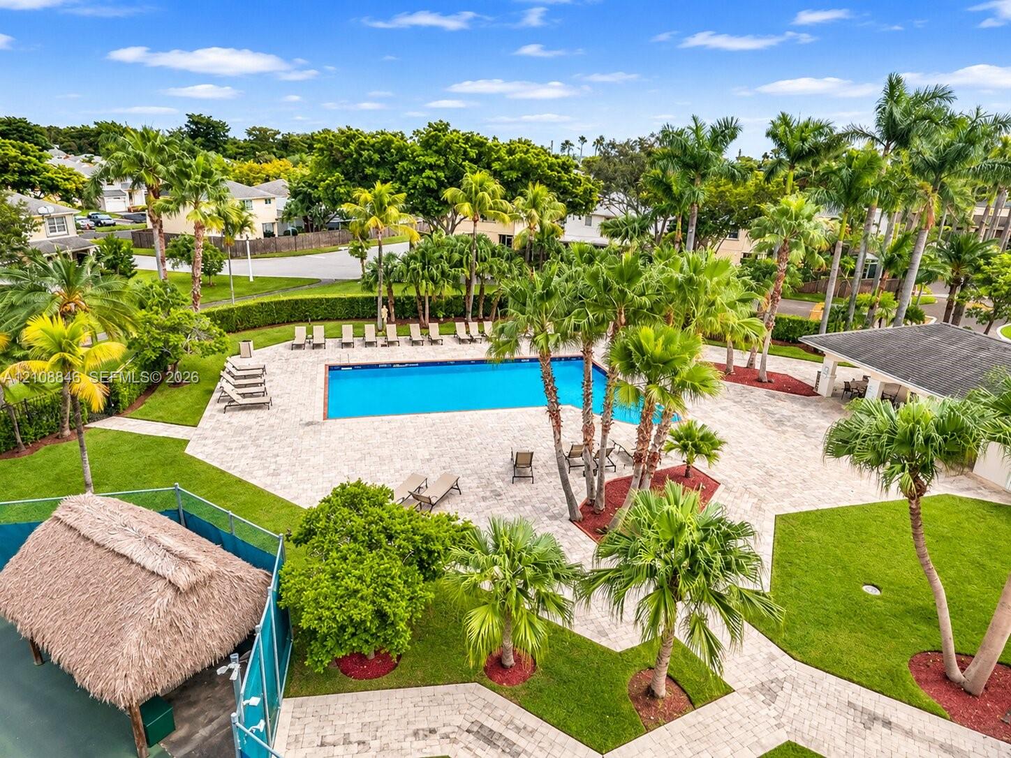 Community swimming pool featuring a paved deck, surrounding mature palm trees, manicured lawns, and a thatched-roof pavilion