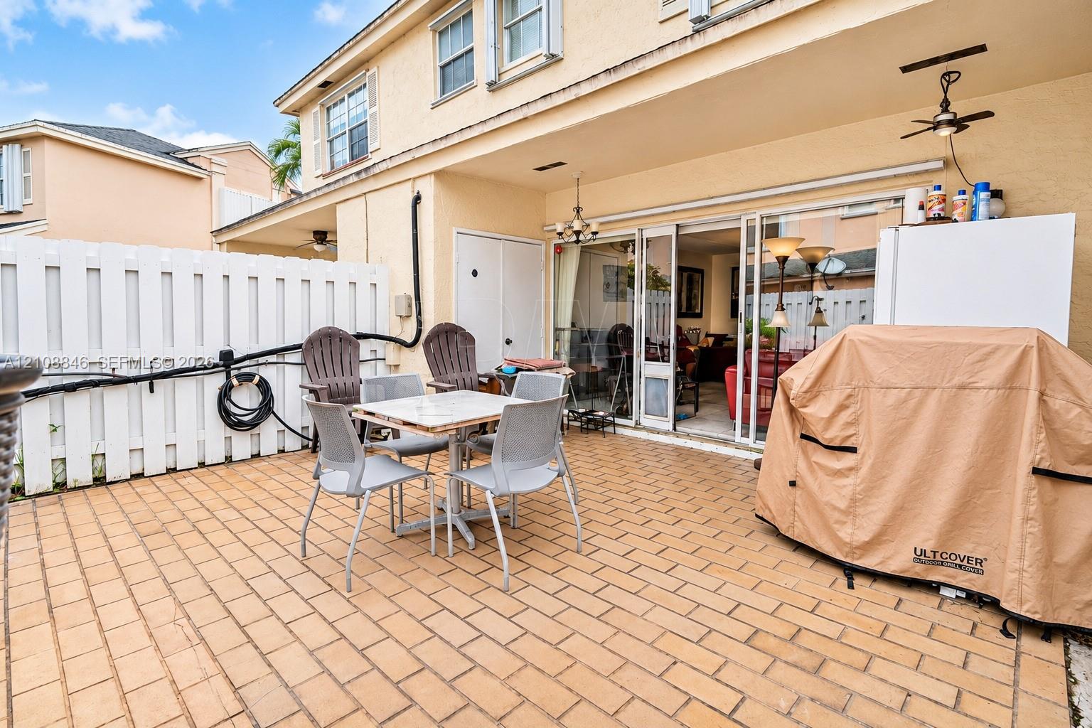 Covered concrete patio with paver-style finish, multi-panel sliding glass doors, white vinyl privacy fencing, and ceiling-mounted light fixtures