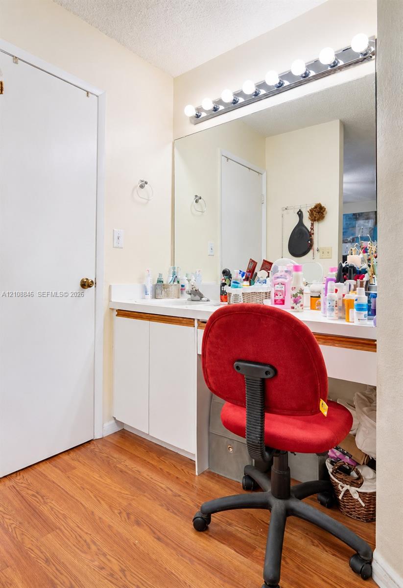 Built-in vanity featuring white cabinetry, a solid surface countertop, an expansive wall-to-wall mirror, integrated multi-bulb vanity lighting, and wood-finish flooring