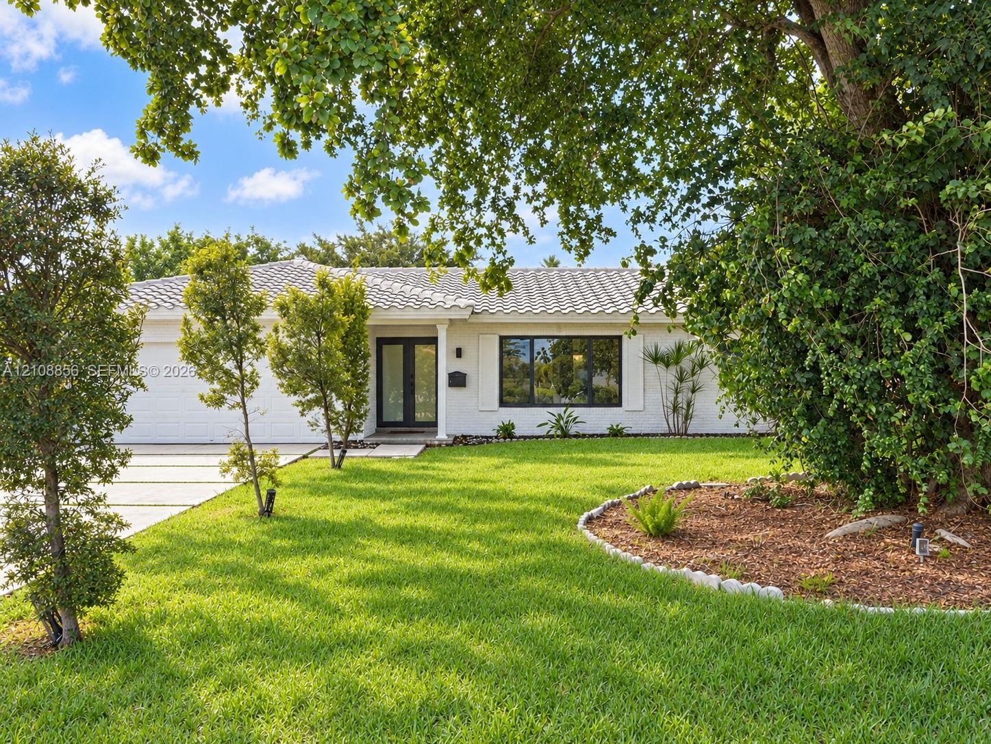 Single-story residence featuring a white painted brick exterior, tile roof, expansive front lawn, landscaped mulch garden beds with stone border, and a concrete driveway