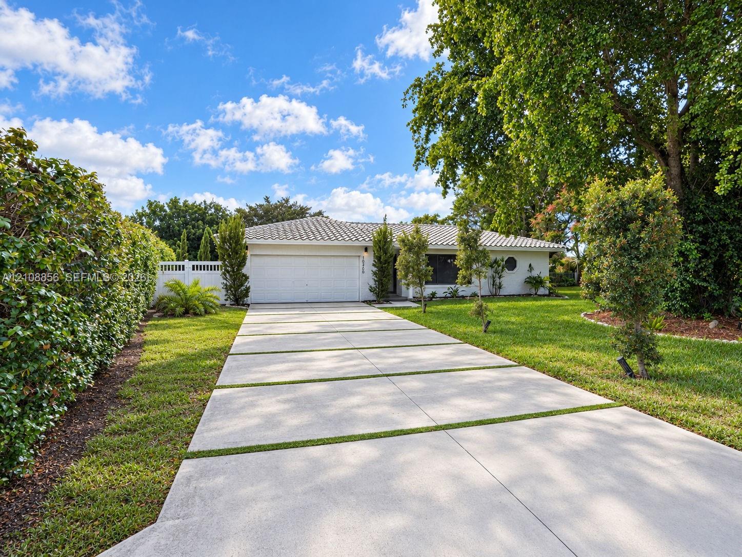 Single-story residence featuring a light-toned stucco exterior, a segmented concrete driveway, a multi-car garage, a tiled roof, and mature perimeter landscaping