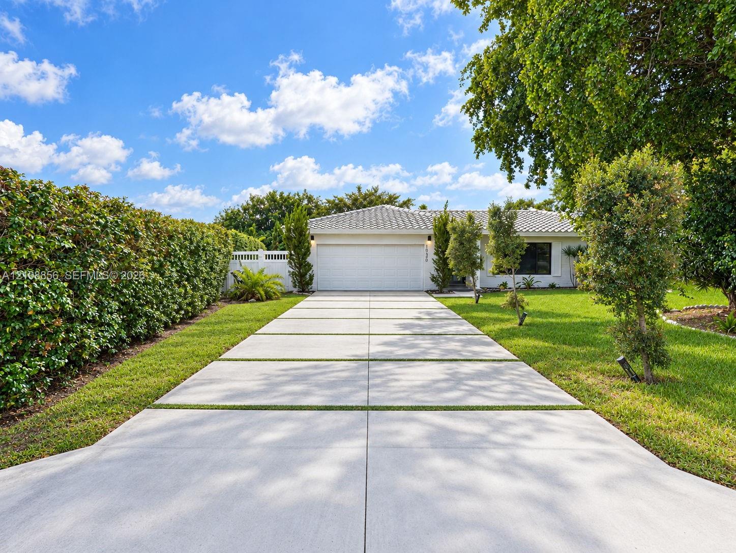 Single-story residence featuring a tile roof, an expansive concrete driveway with grass inlays, a wide garage door, and lush manicured hedges