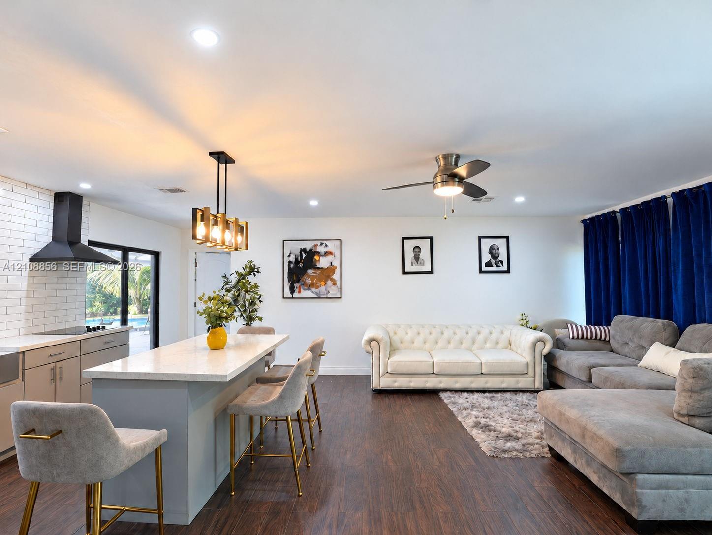 Open-concept kitchen and living area featuring a center island with light-toned solid surface countertop, white brick backsplash, matte black range hood, wood-finish flooring, and recessed ceiling lighting