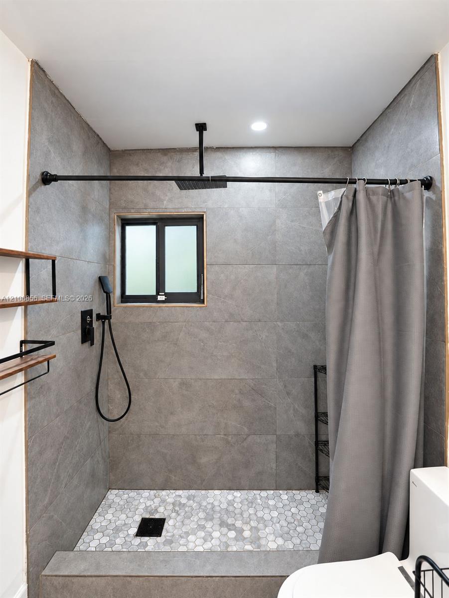 Walk-in shower featuring large-format grey tile surround, hexagonal floor tiling, a ceiling-mounted rainfall showerhead, and matte black plumbing fixtures