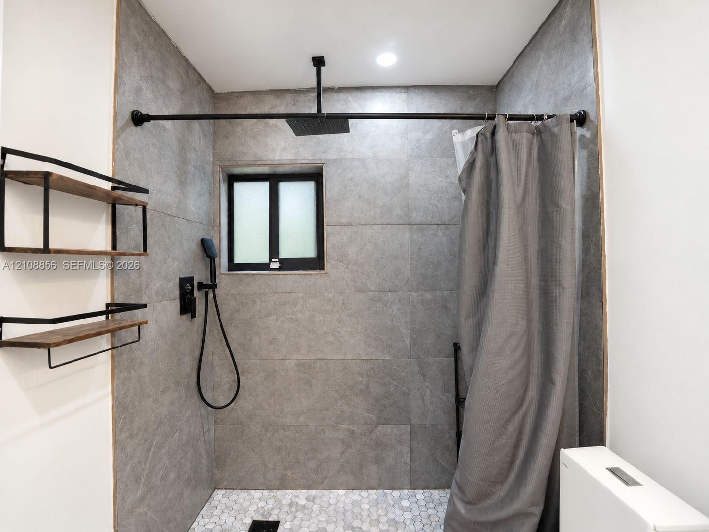 Shower area featuring large-format grey tile walls, a white hexagonal tile floor, matte black rainfall showerhead with handheld sprayer, and built-in wood-finish shelving with black metal framing