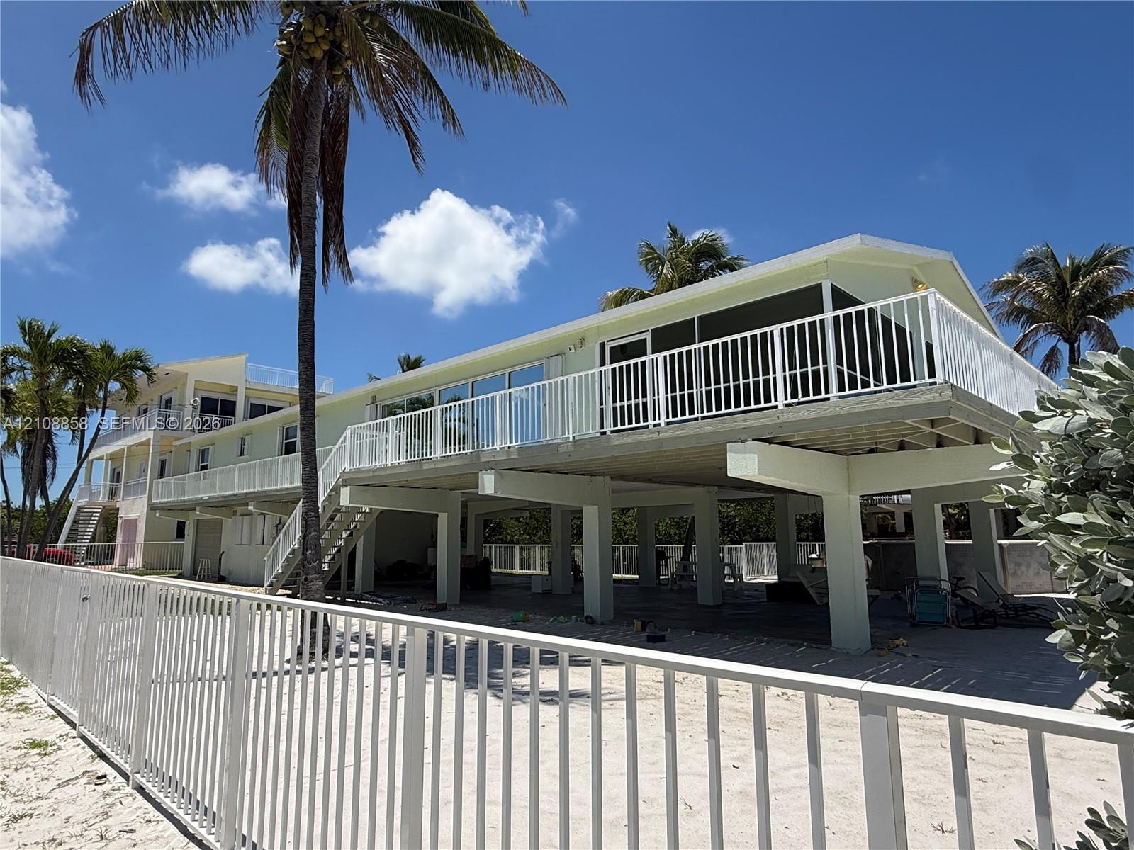 Elevated exterior featuring white metal railings, a screened balcony, concrete pilings, and a sandy ground-level area Elevated exterior featuring white metal railings, a screened balcony, concrete pilings, and a sandy ground-level area