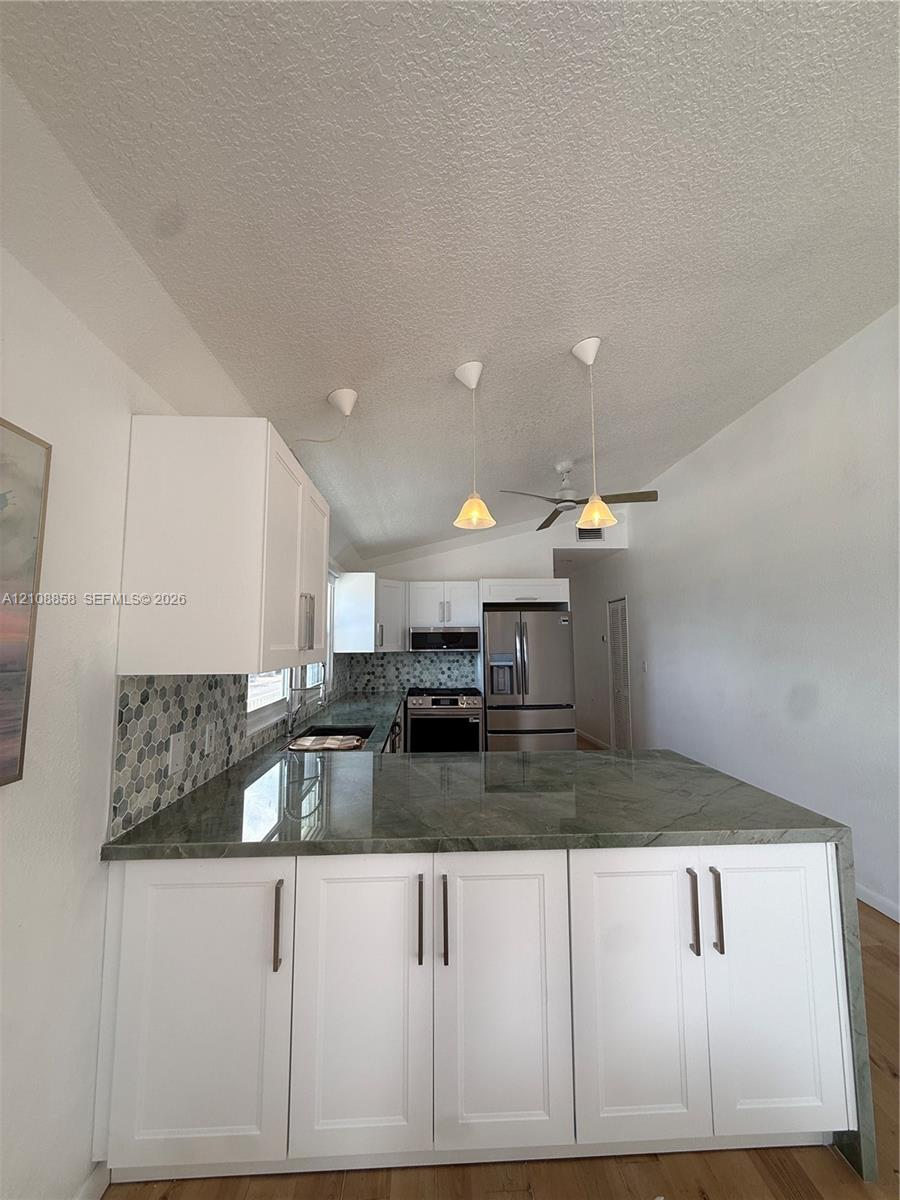 Kitchen featuring white shaker-style cabinetry, solid-surface countertops with waterfall edge, geometric tile backsplash, stainless steel appliances, and pendant lighting
