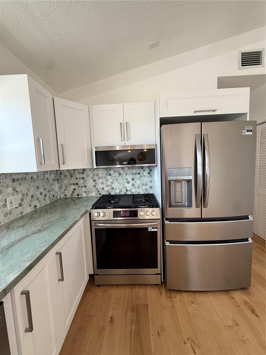 Kitchen featuring white shaker-style cabinetry, natural stone countertops, hexagonal tile backsplash, stainless steel appliances, and wood-finish flooring