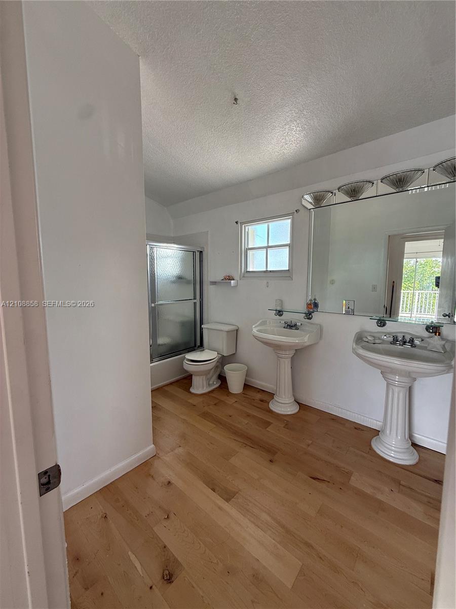 Bathroom featuring wood-finish flooring, dual pedestal sinks, a large wall-mounted mirror with integrated lighting, and a glass-enclosed shower