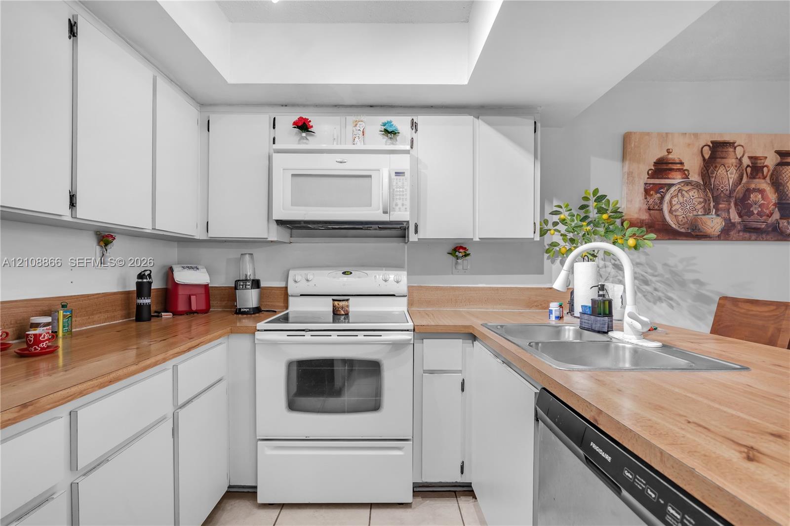 Kitchen featuring white shaker-style cabinetry, wood-finish countertops, tile flooring, a dual-basin stainless steel sink with a high-arch faucet, and an integrated white microwave