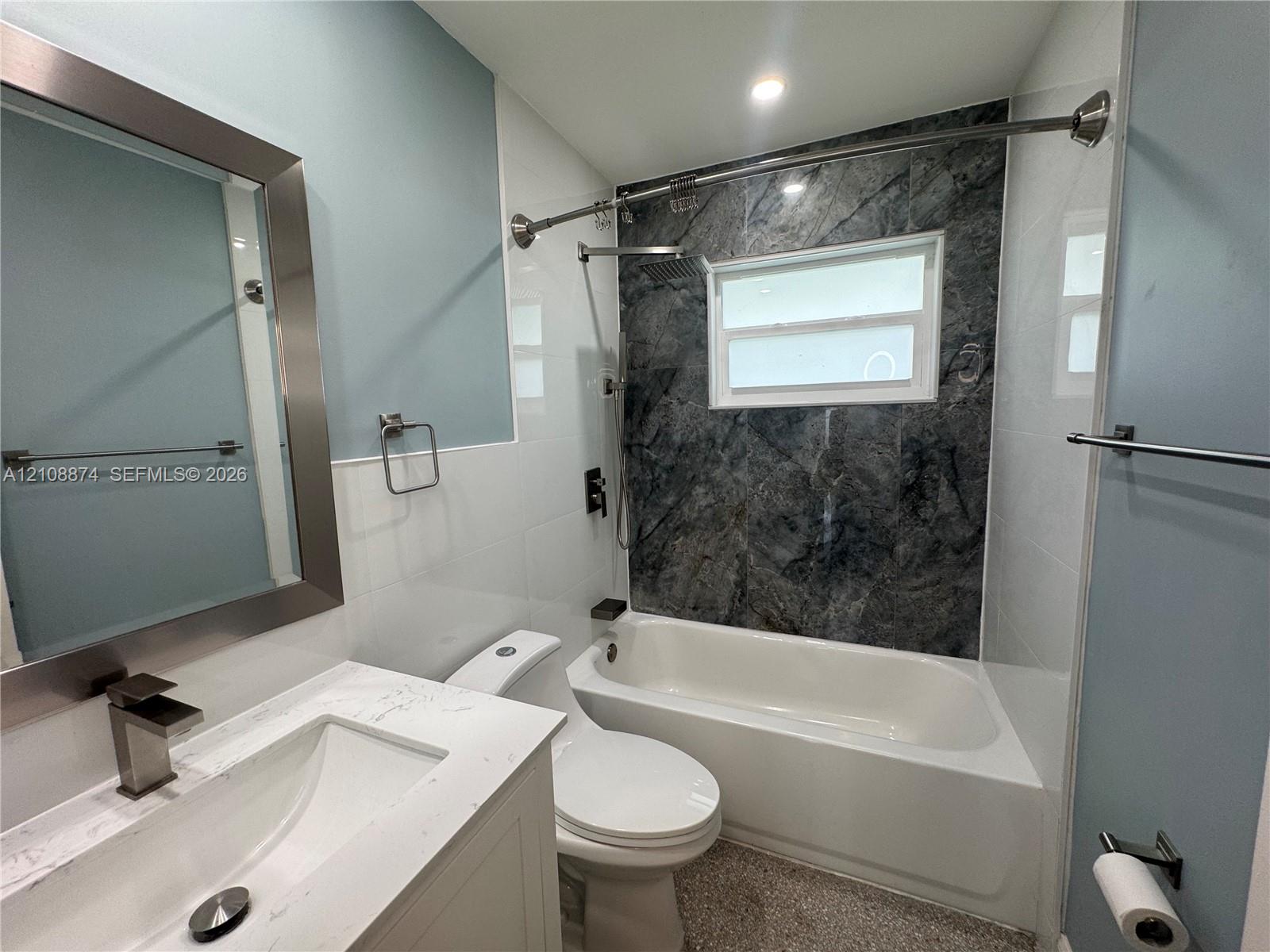 Bathroom featuring a white vanity with a stone-top integrated sink, a deep soaking tub with a patterned tile surround, and a framed mirror with metallic finish