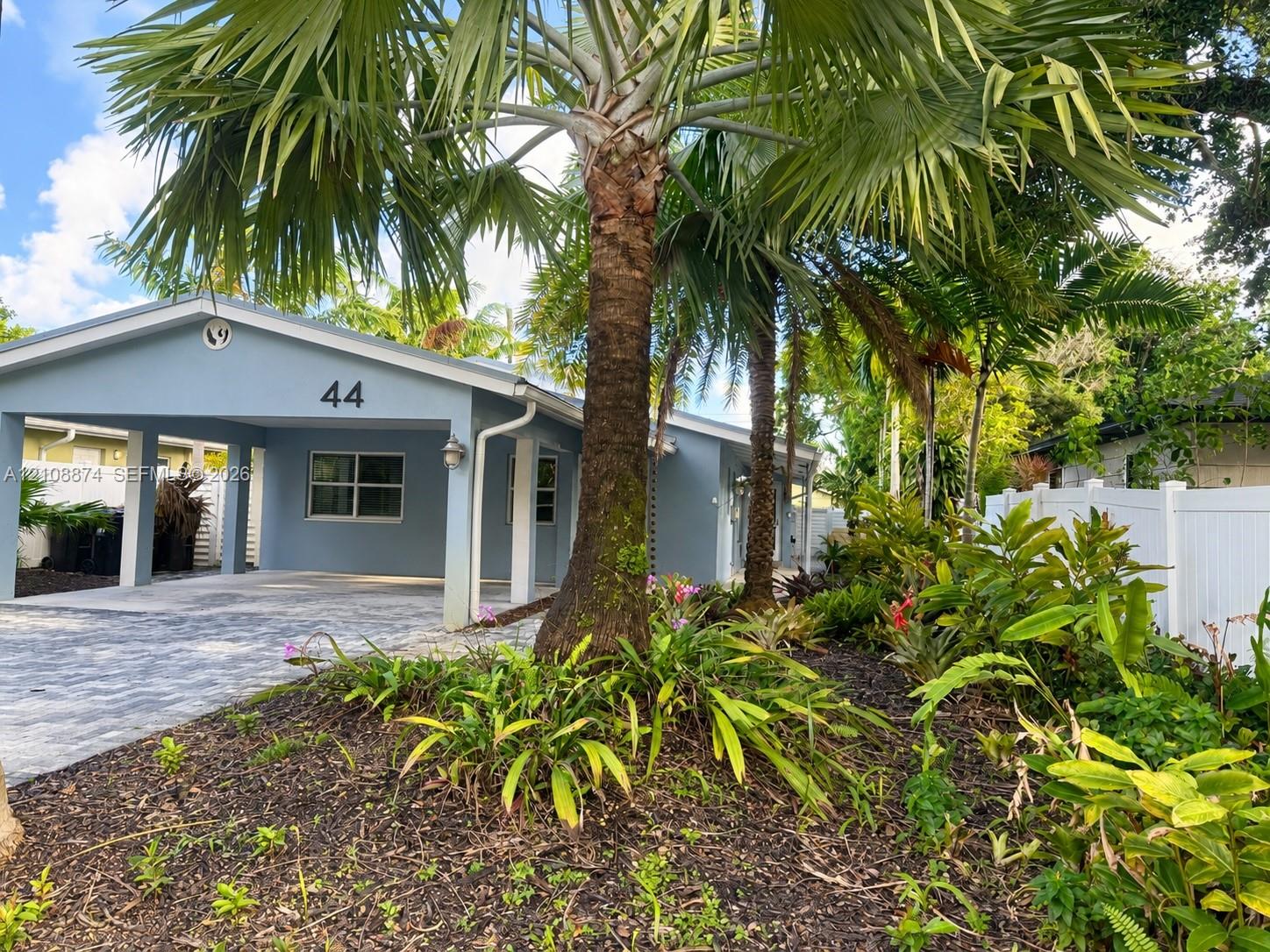 Exterior featuring a covered carport, paver driveway, mature palm trees, and white vinyl privacy fencing