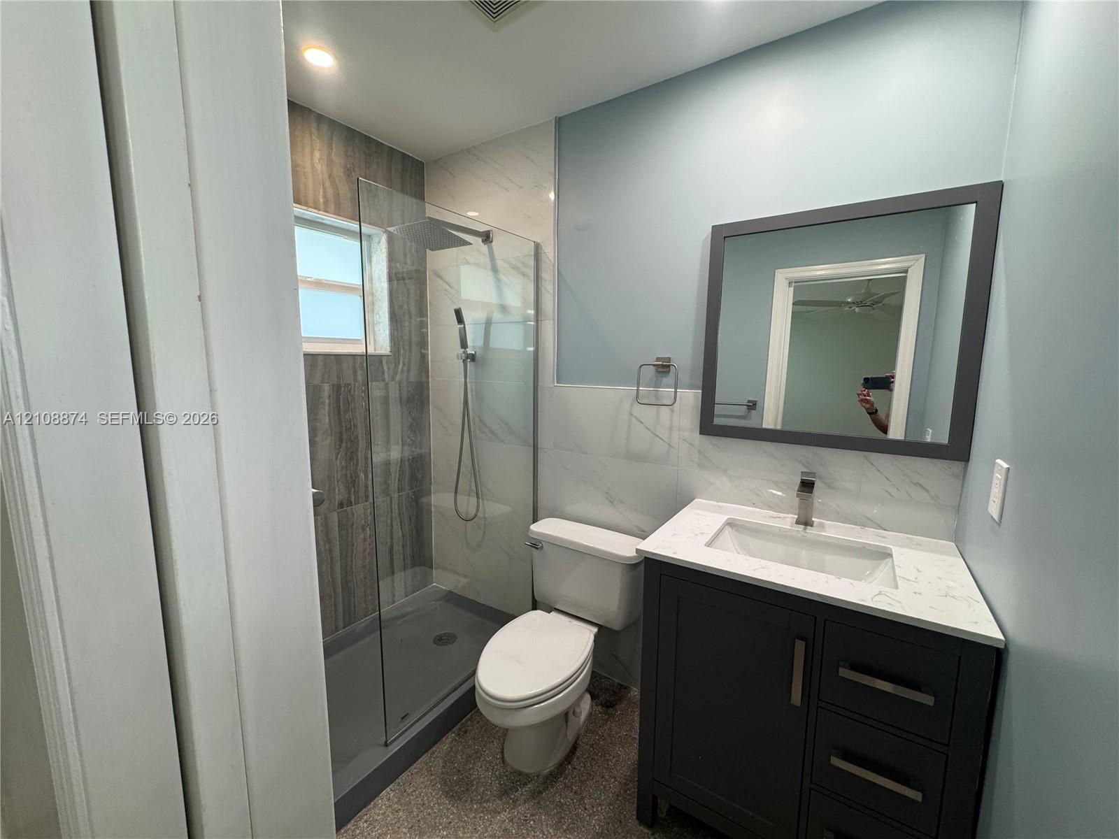 Bathroom featuring a dark vanity with a white countertop and rectangular undermount sink, walk-in shower with floor-to-ceiling tile, glass enclosure, and a wall-mounted rectangular mirror