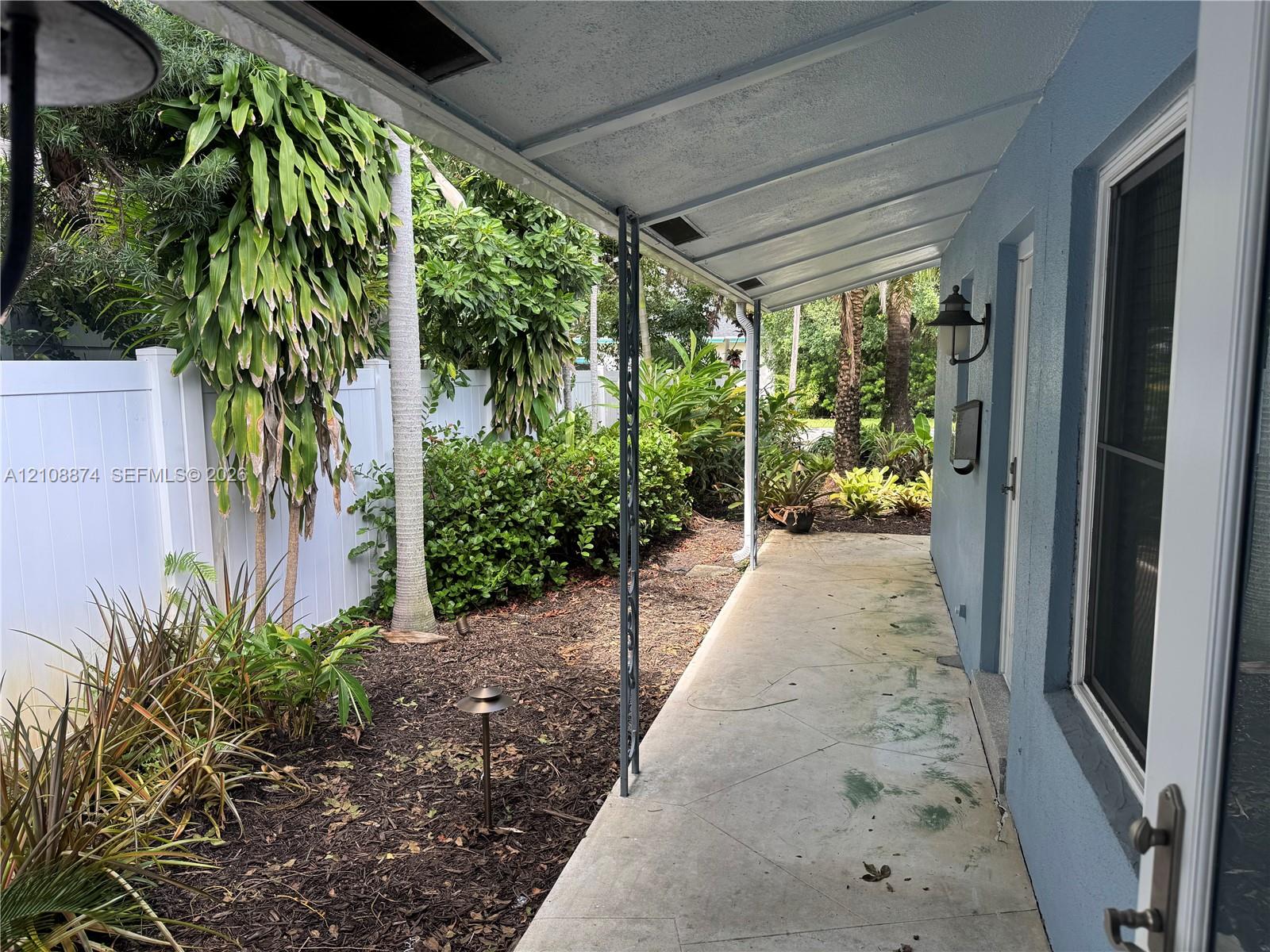 Covered concrete patio featuring decorative metal support columns, a white vinyl privacy fence, and integrated landscape lighting