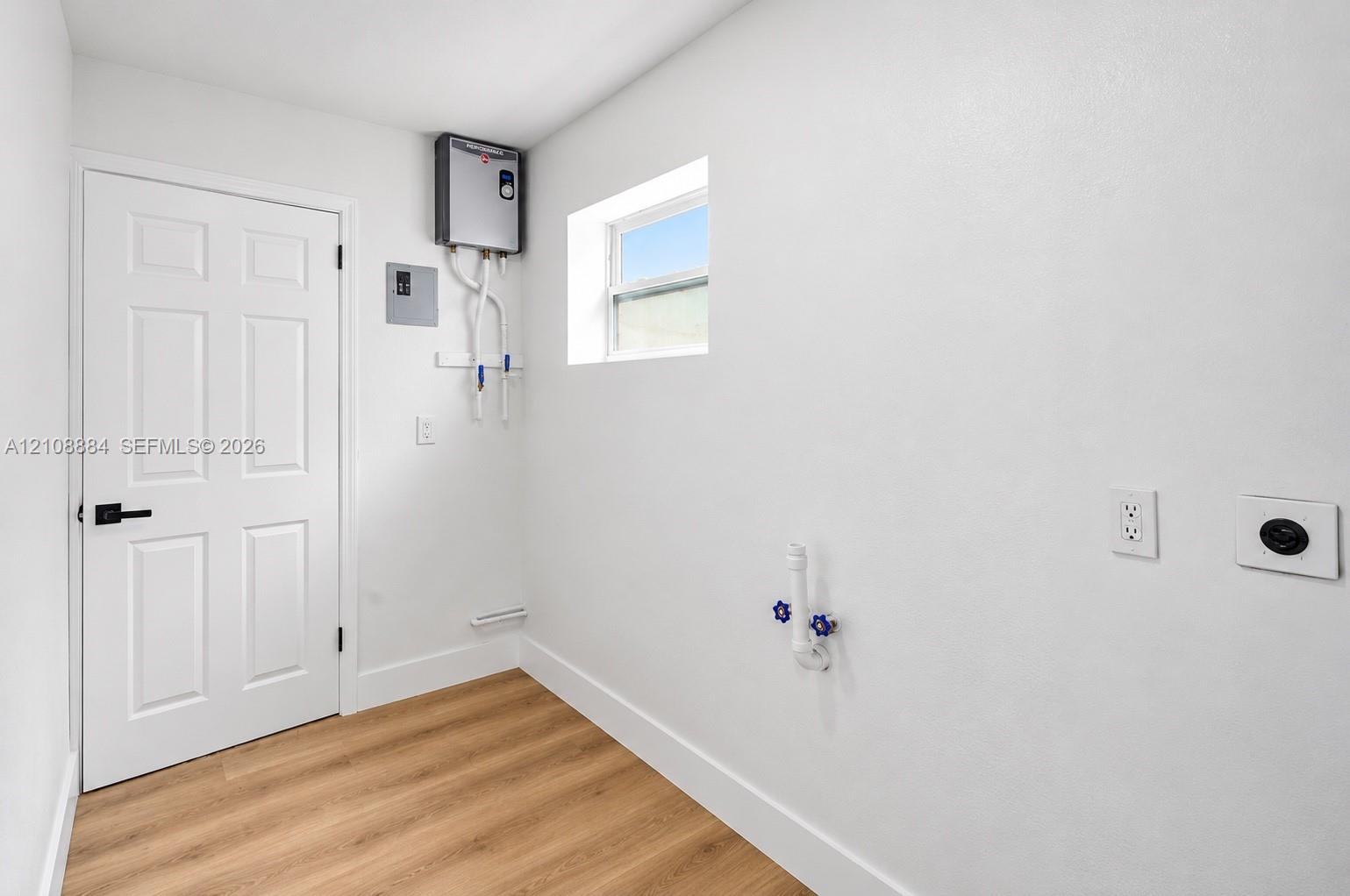 Utility space featuring wood-finish flooring, a wall-mounted tankless water heater, white six-panel door with matte black hardware, and an egress window