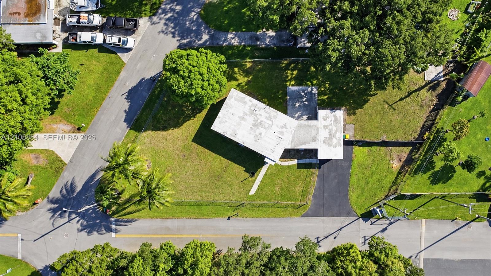 Aerial view of a rectangular property featuring a paved asphalt driveway, expansive green lawn, mature trees, and a flat-roof residence
