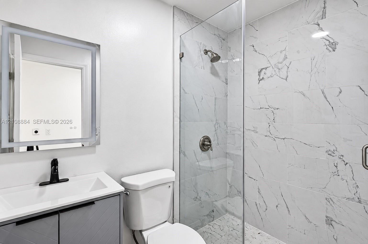 Bathroom featuring a gray vanity with a white countertop, wall-mounted LED-backlit mirror, and walk-in shower with floor-to-ceiling tile surround and glass enclosure