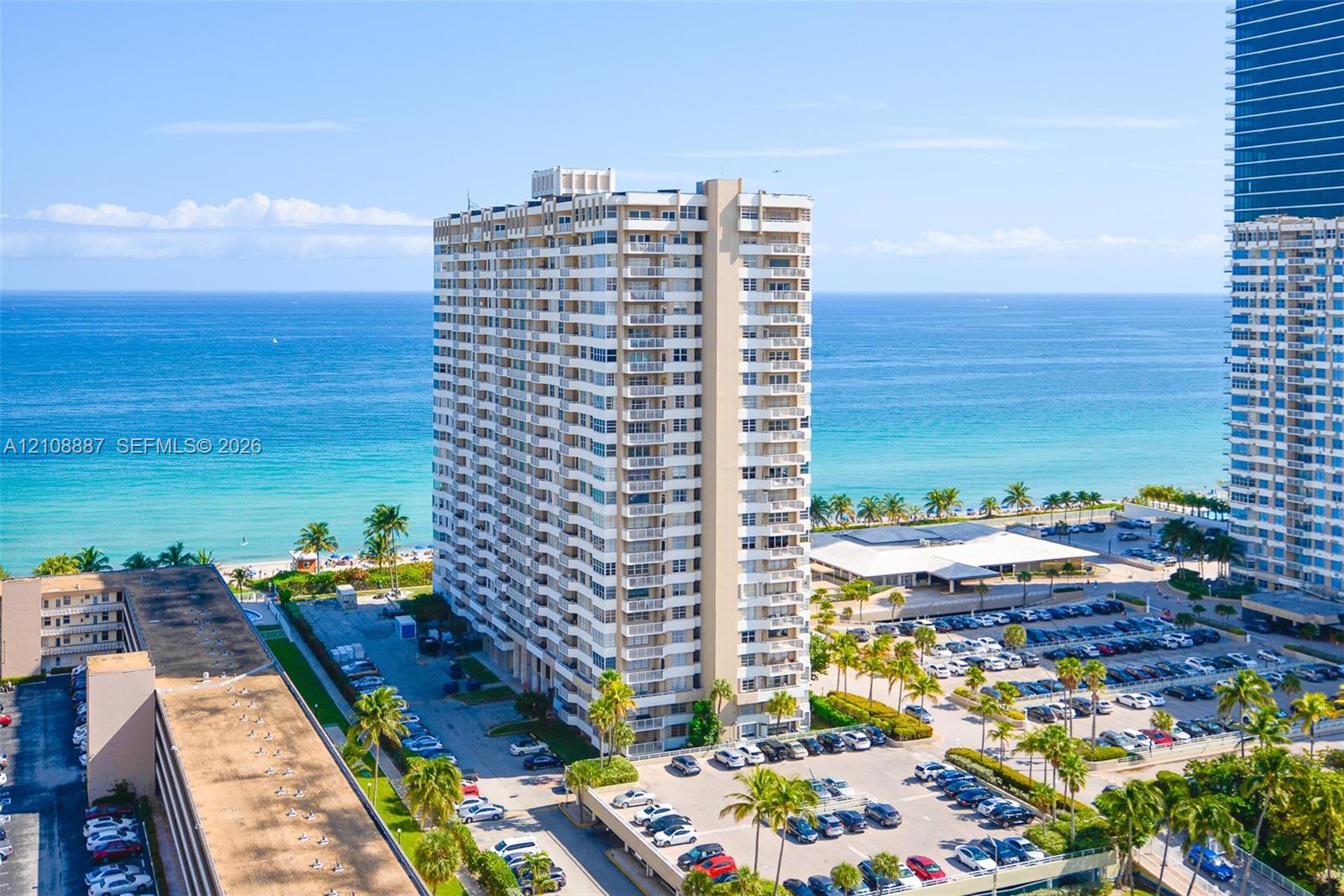 High-rise residential building featuring stacked private balconies, an expansive paved parking area, and mature palm tree landscaping High-rise residential building featuring stacked private balconies, an expansive paved parking area, and mature palm tree landscaping