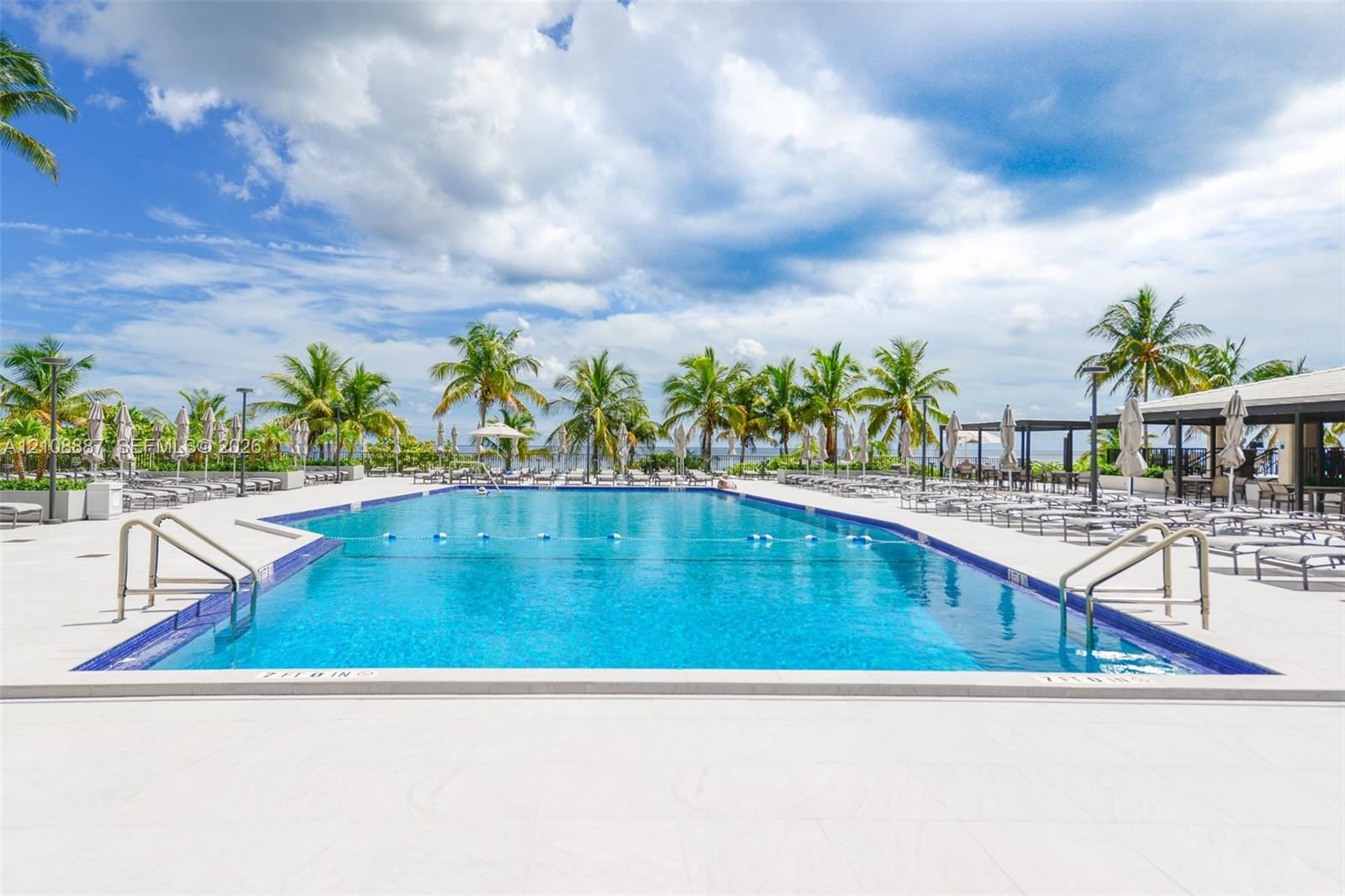 Large swimming pool with tiled perimeter, metal handrails, and a wide paved deck surrounded by mature palm trees