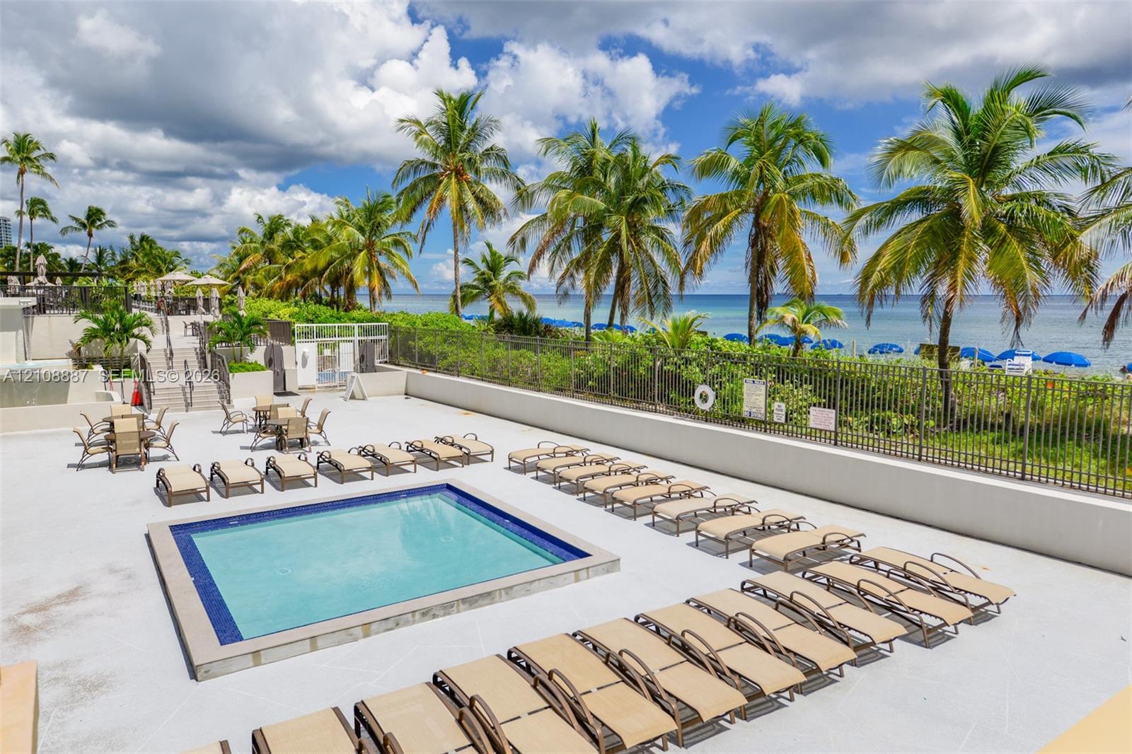 Swimming pool with tiled border, expansive paved patio, and perimeter metal fencing overlooking the oceanfront
