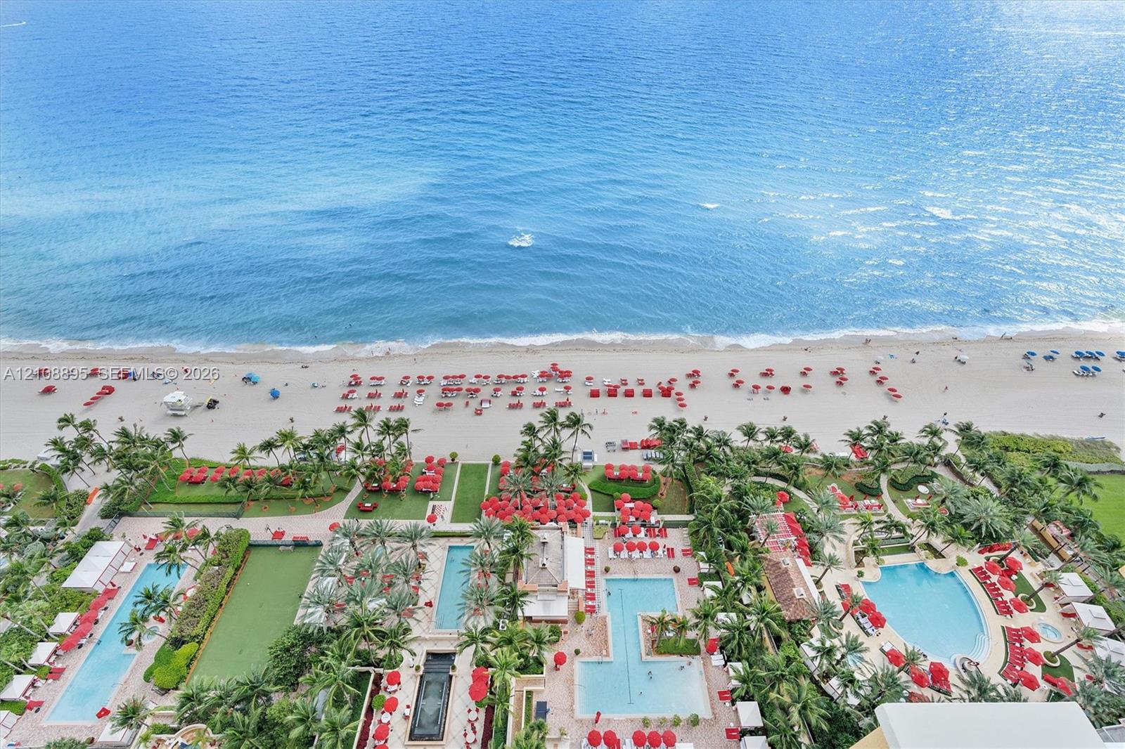 Aerial perspective of oceanfront property featuring multiple swimming pools, expansive paved sun decks, manicured palm landscaping, and direct access to the beach