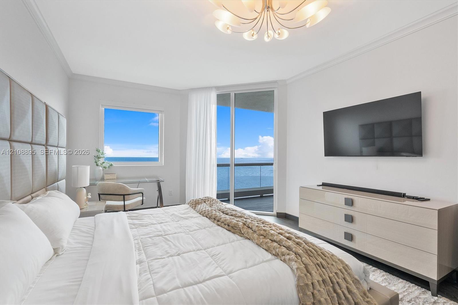 Spacious interior featuring a decorative multi-arm chandelier, ceiling crown molding, floor-to-ceiling glass sliding door, private balcony, and window with ocean backdrop
