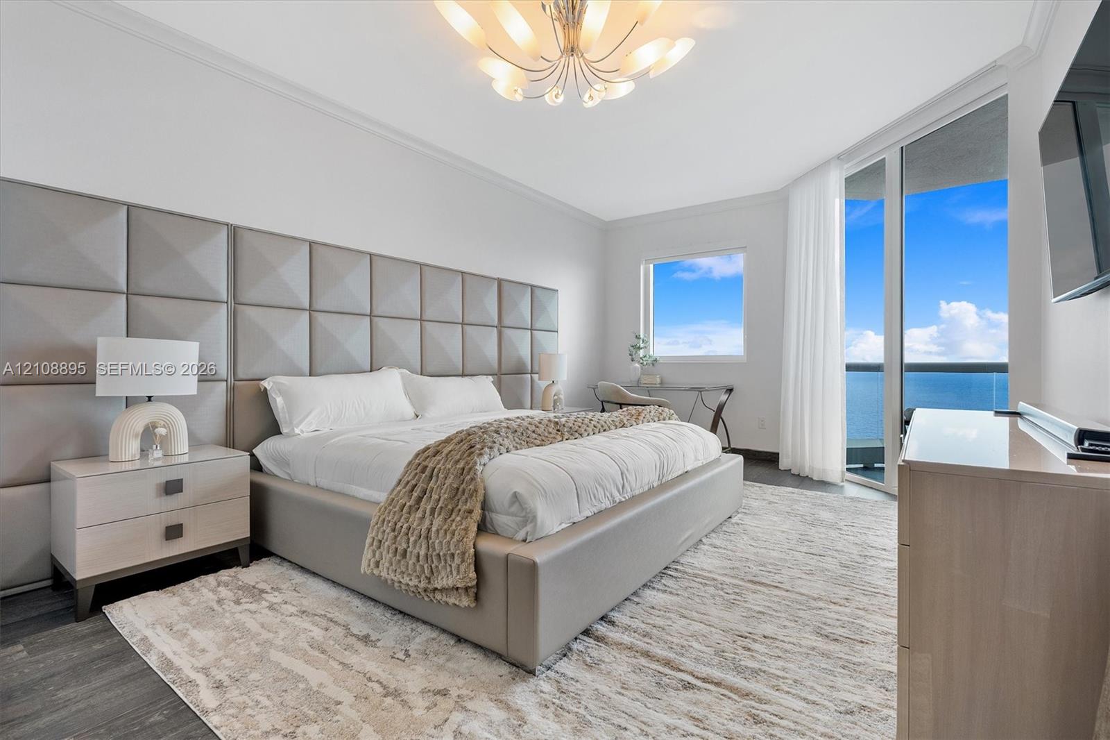 Floor-to-ceiling glass door opening to a private balcony, custom upholstered wall paneling, multi-arm chandelier, and dark wood-finish flooring