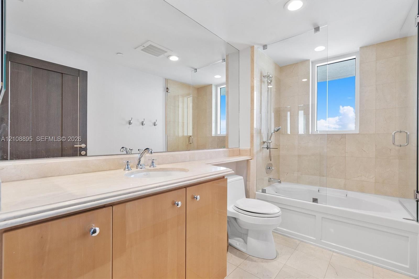 Wood-finish cabinetry with a stone-surface countertop, undermount sink, expansive vanity mirror, and a tiled shower-tub combination featuring a glass enclosure