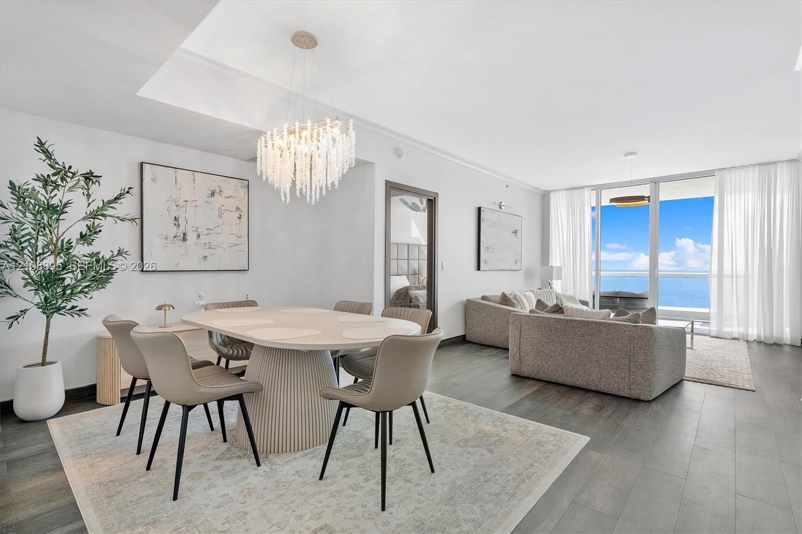 Open-concept living space featuring wood-finish flooring, a contemporary multi-tiered crystal chandelier, recessed ceiling lighting, and floor-to-ceiling glass paneling with an ocean backdrop