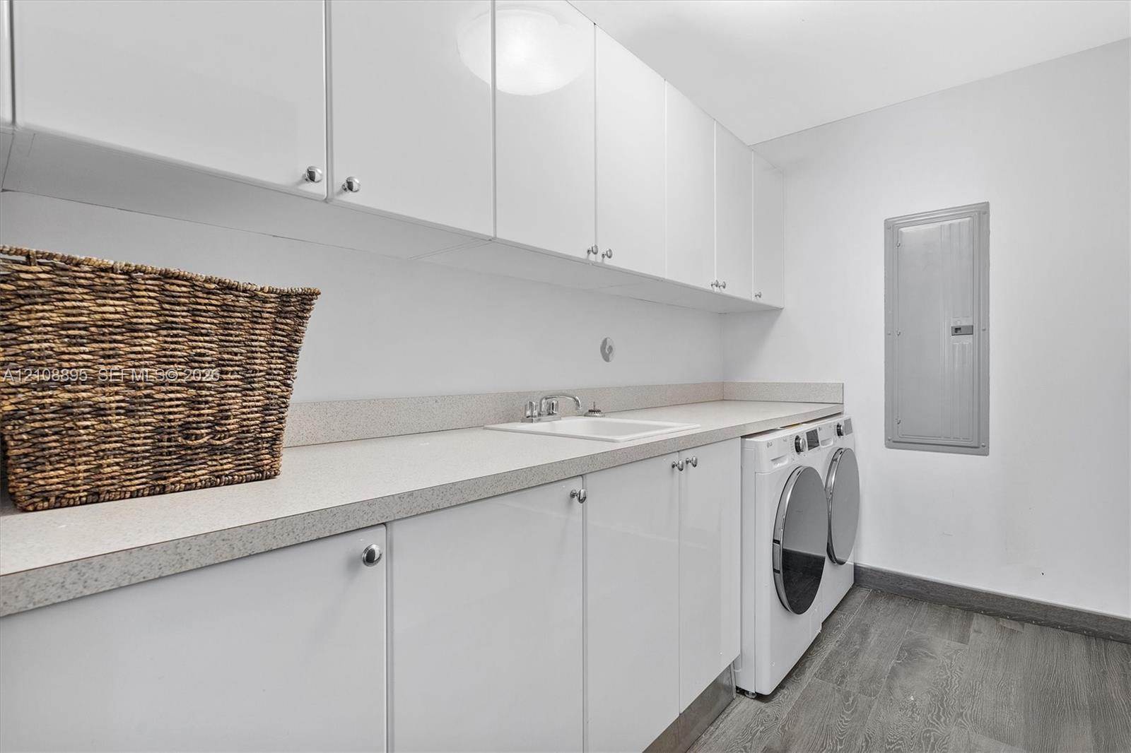 Laundry area featuring high-gloss white upper and lower cabinetry, stone-composite countertop with an integrated sink, metal faucet hardware, and wood-finish flooring