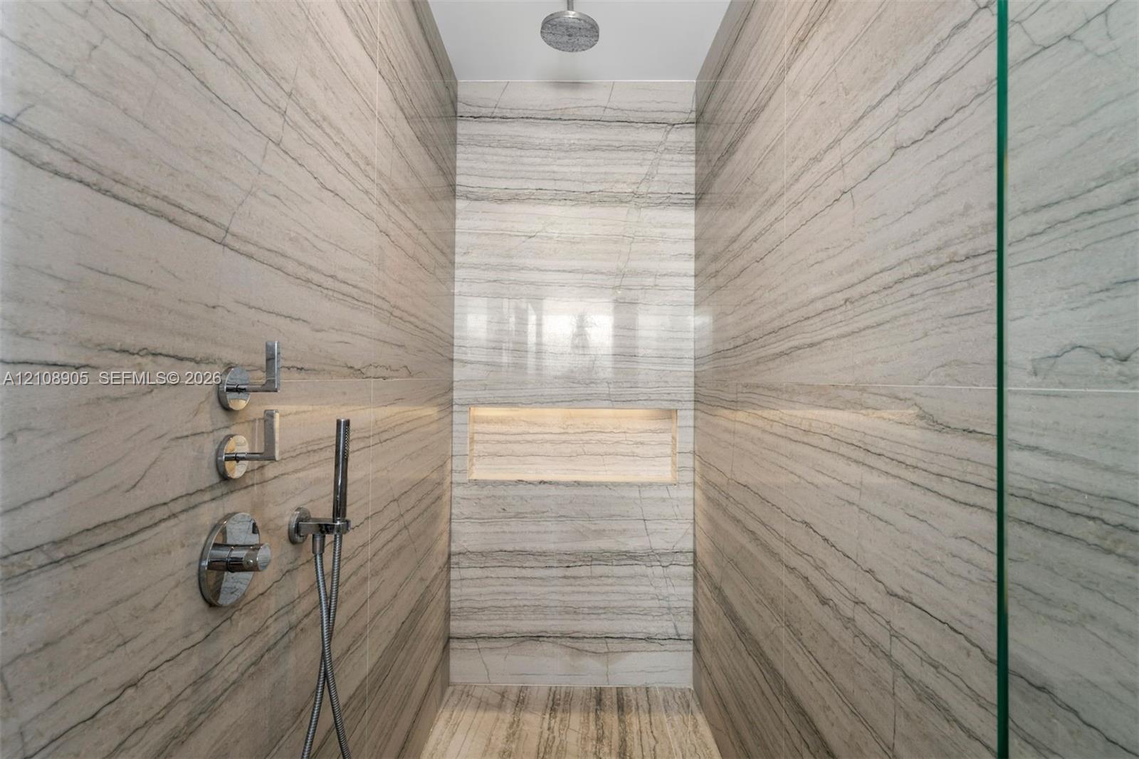 Walk-in shower featuring floor-to-ceiling stone-tiled walls, a built-in recessed niche, chrome wall-mounted fixtures, a handheld sprayer, and a ceiling-mounted rainfall showerhead
