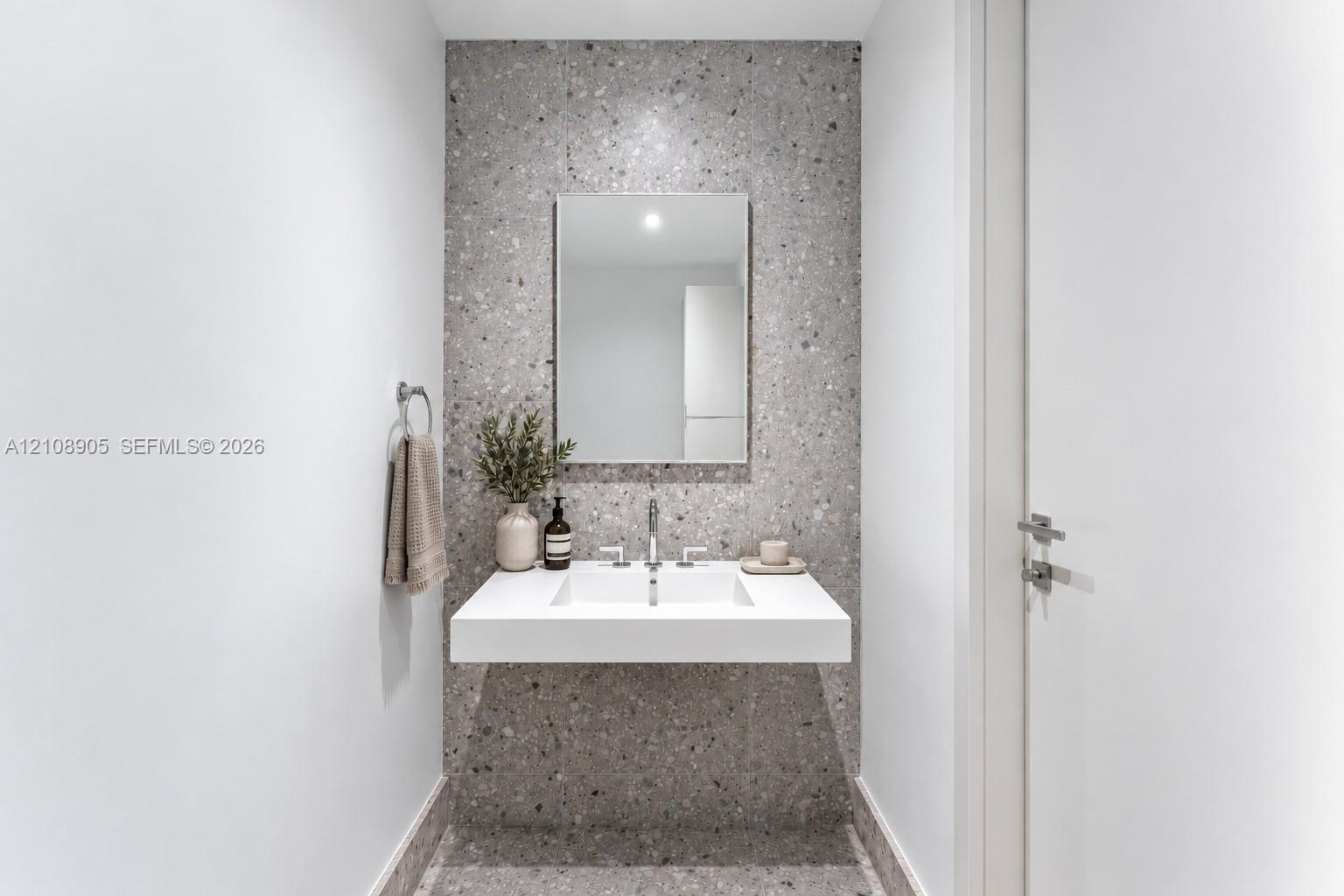 Floating vanity with a rectangular integrated sink, polished metal widespread faucet, rectangular wall-mounted mirror, and speckled stone-finish wall cladding