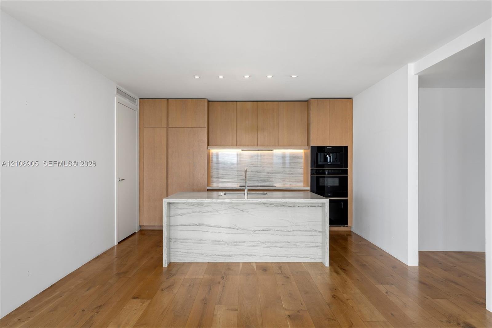 Modern kitchen featuring custom wood-finish cabinetry, a waterfall-edge stone island, built-in dual ovens, integrated cooktop, and wood-finish flooring
