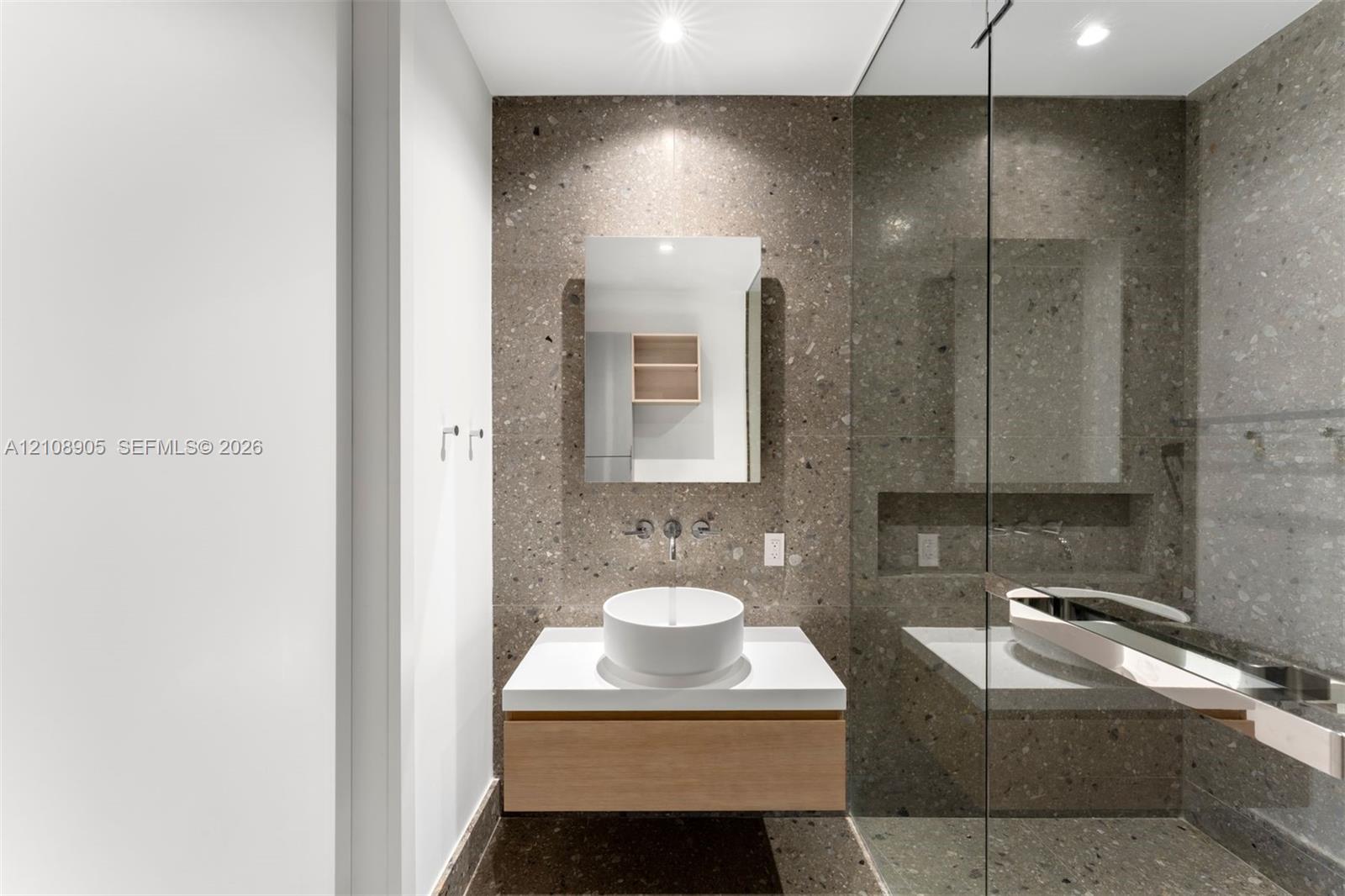 Floating vanity with a white vessel sink, wall-mounted faucet, rectangular mirror, glass-enclosed shower, and large-format stone-look wall and floor tiling
