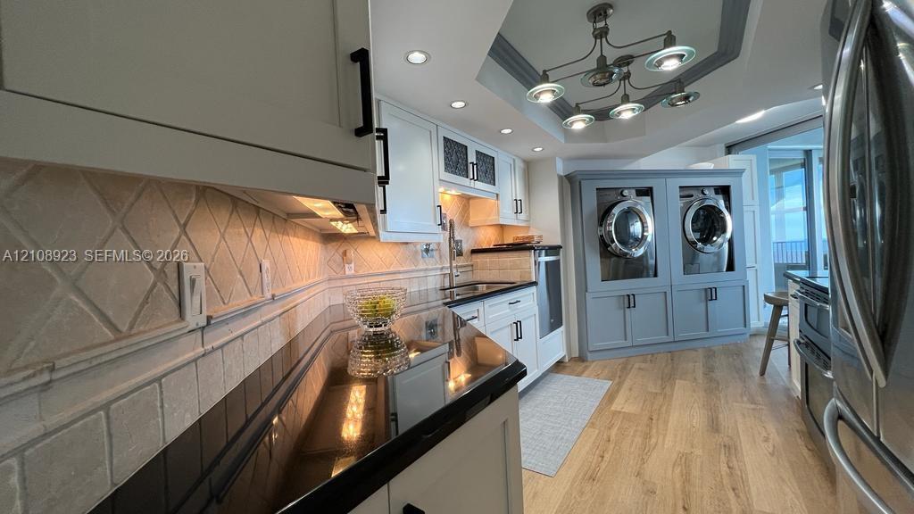Kitchen featuring white shaker cabinetry, dark solid-surface countertops, diamond-patterned tile backsplash, wood-finish flooring, and a tray ceiling with integrated lighting