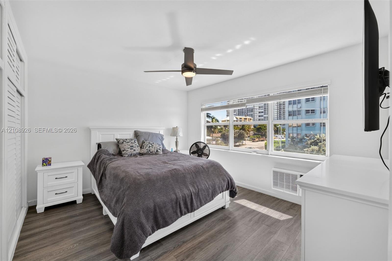 Room featuring wood-finish flooring, louvered-style built-in closet doors, a multi-pane window, and a ceiling-mounted light fixture with fan