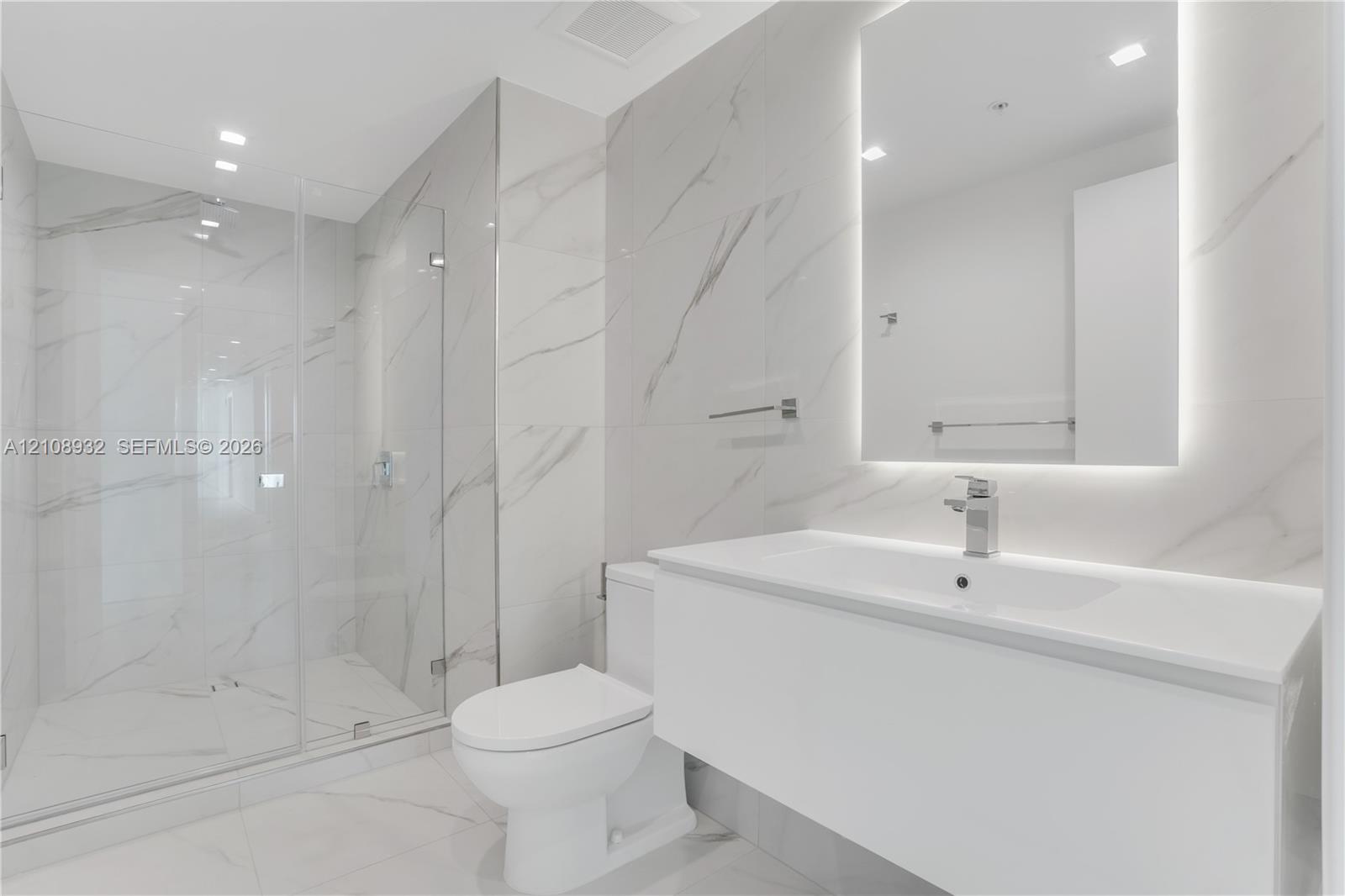 Modern bathroom featuring floor-to-ceiling large-format stone-patterned tile, a glass-enclosed walk-in shower, a white floating vanity with a seamless countertop, and a wall-mounted mirror with integrated backlighting